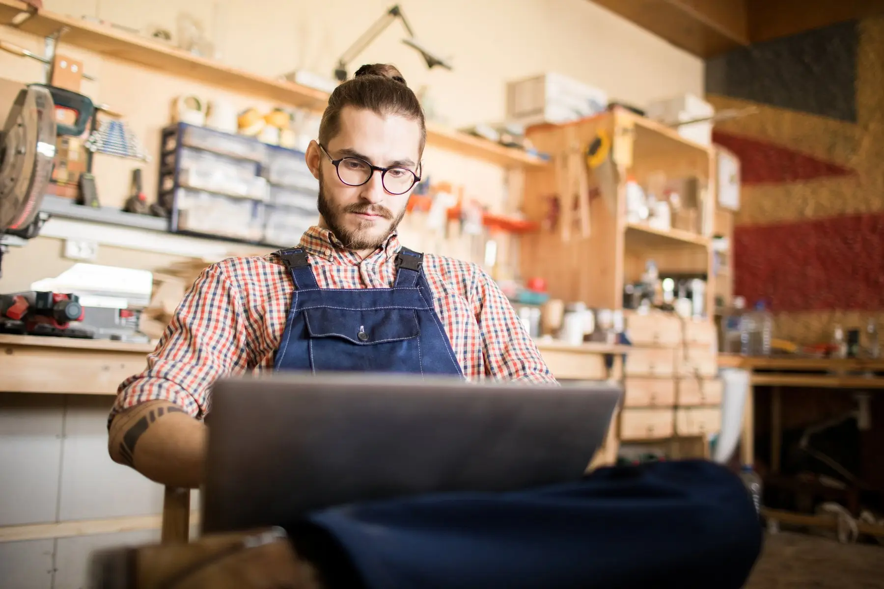 Un artisan en salopette et chemise à carreaux travaille sur son ordinateur portable dans son atelier de menuiserie