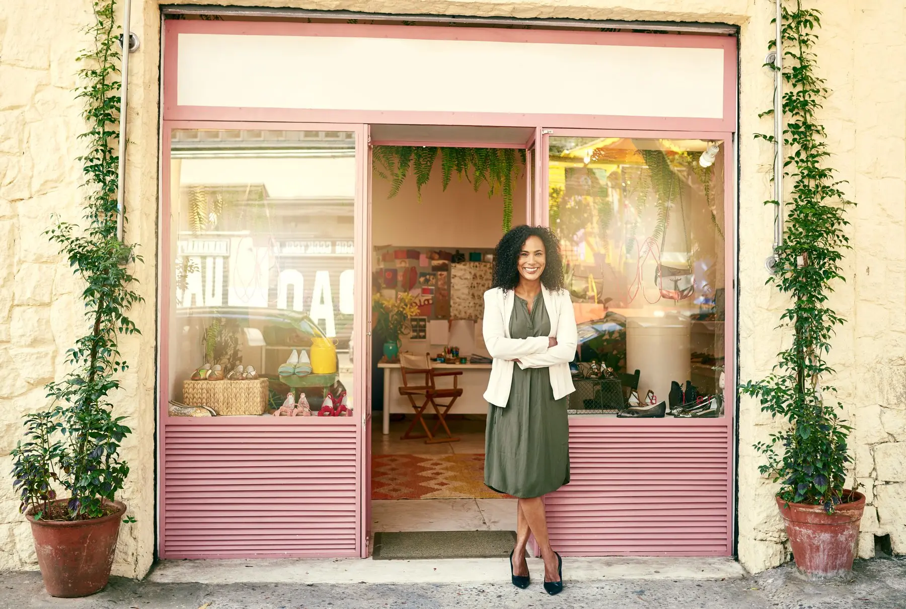 Une commerçante souriante, bras croisés, pose devant l’entrée de sa boutique aux huisseries roses, sous un bandeau d’enseigne encore vierge