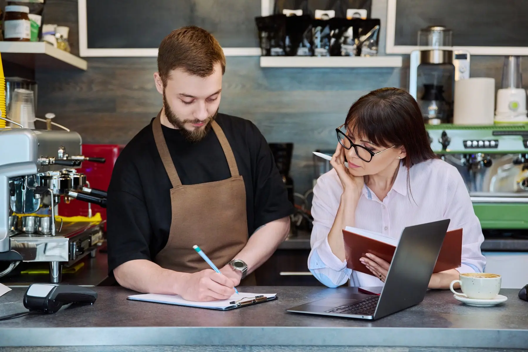Deux commerçants, derrière le comptoir d’un café, comparent leurs notes devant un ordinateur portable