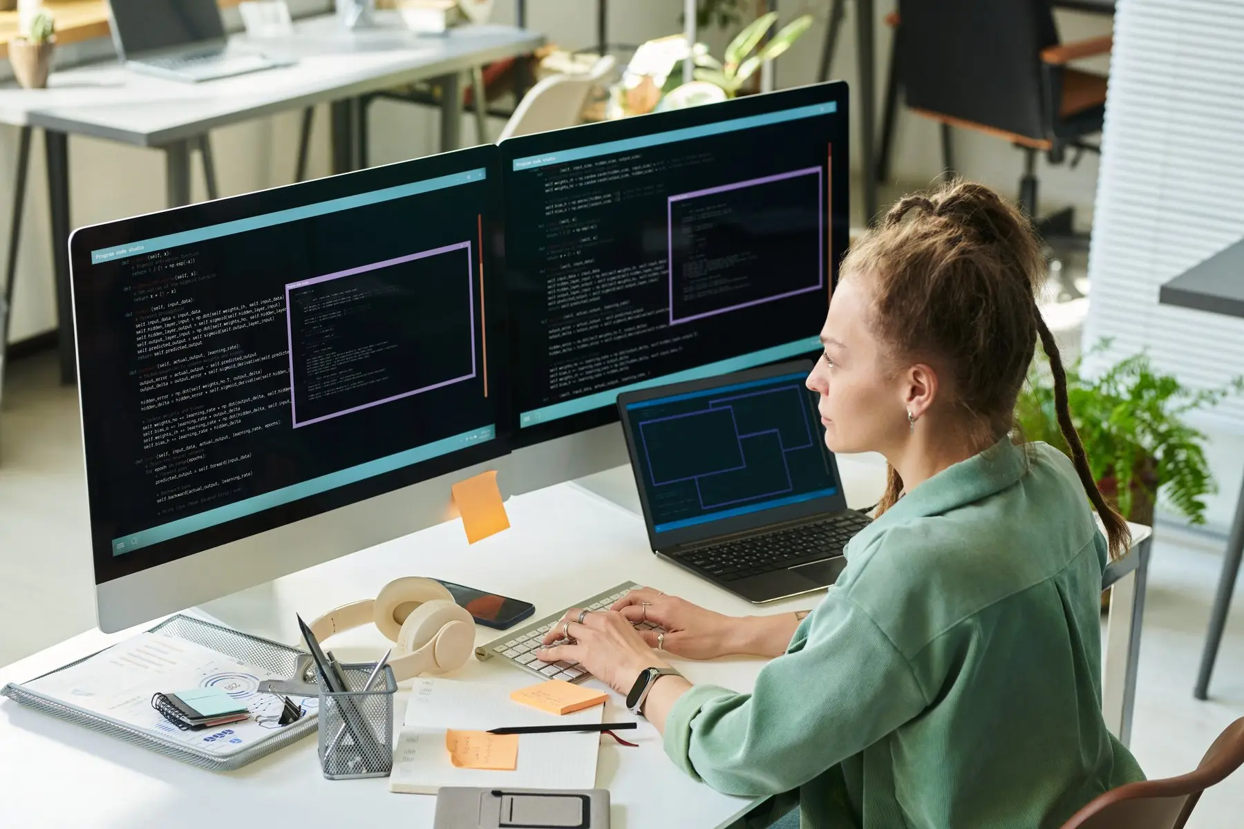Une développeuse travaille face à deux écrans remplis de code, un ordinateur portable ouvert à côté d’elle