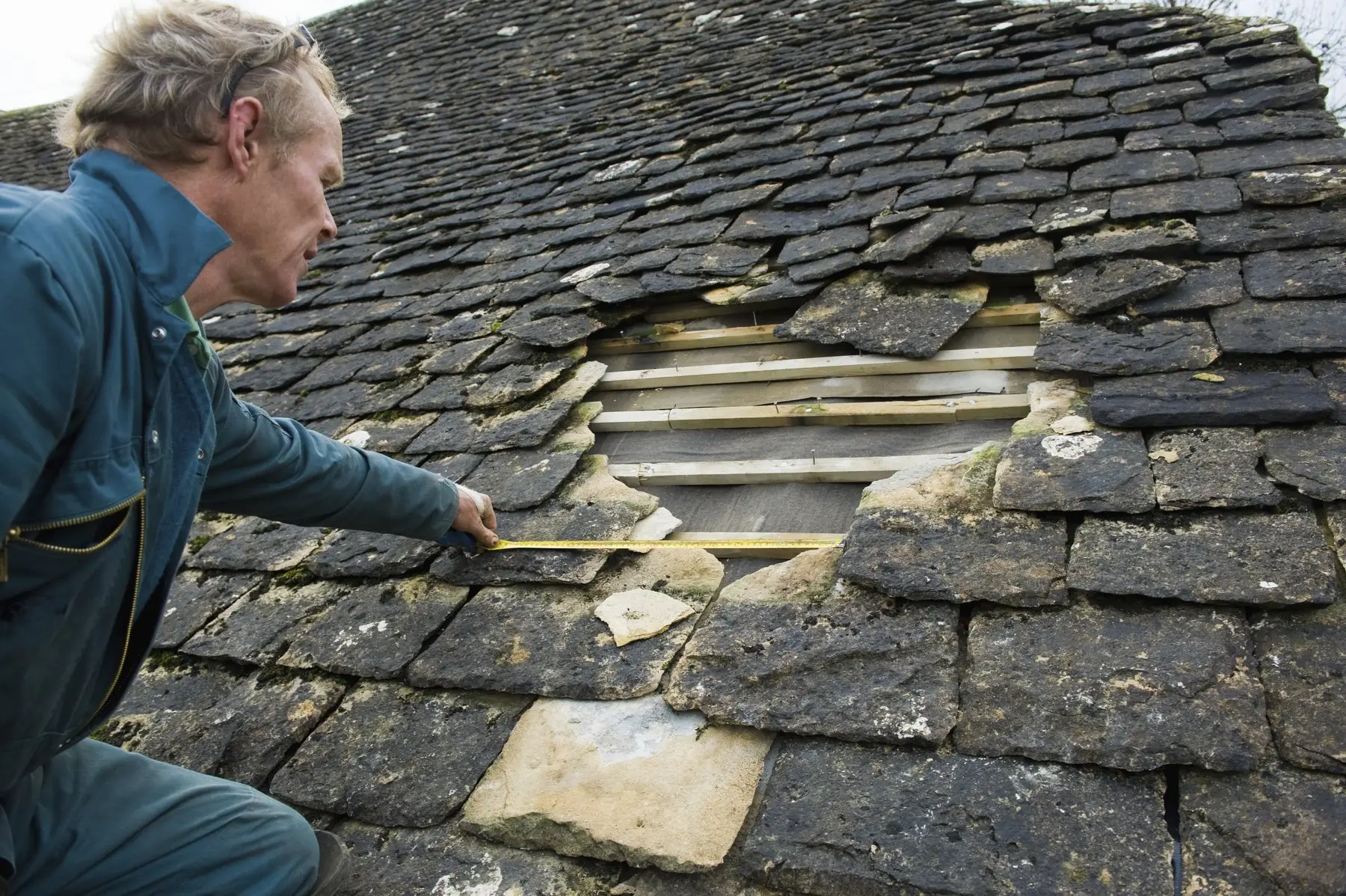 Un couvreur mesure au mètre ruban une réparation sur une toiture en ardoises anciennes