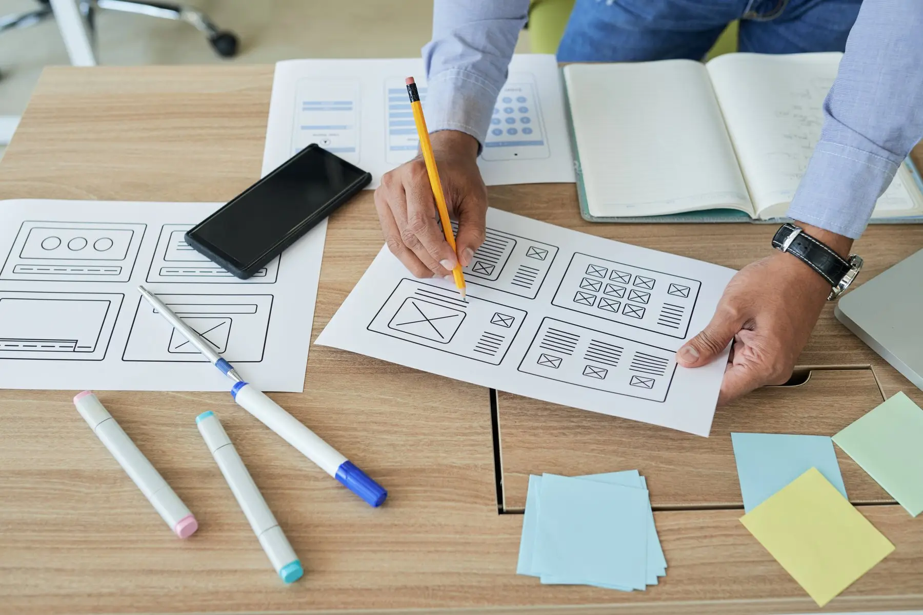 Deux mains dessinent au crayon des croquis de pages web, à côté d’un smartphone, de feutres et de notes adhésives