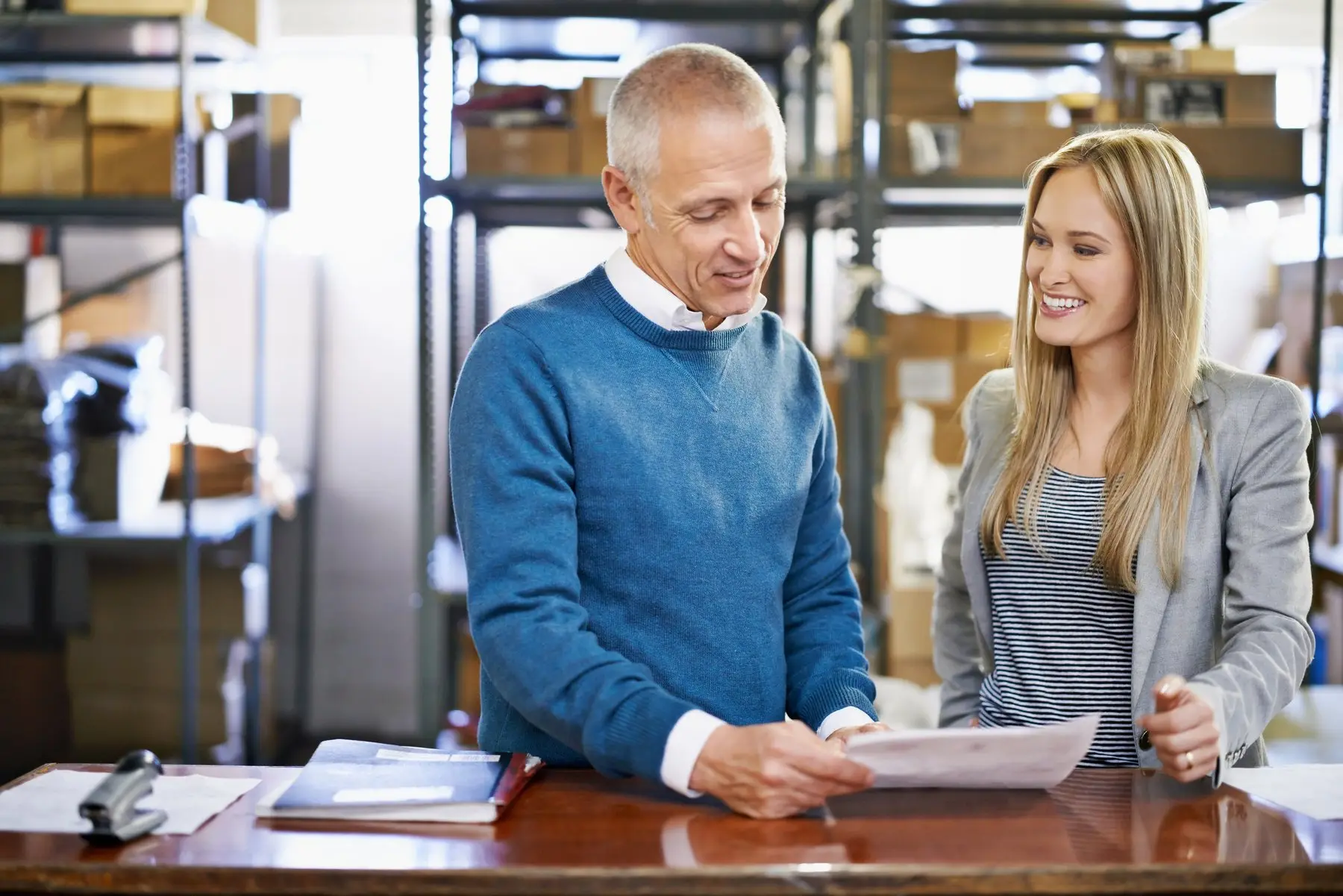 Un homme en pull bleu et une femme en veste grise relisent un document au comptoir d’une réserve, devant des étagères chargées de cartons