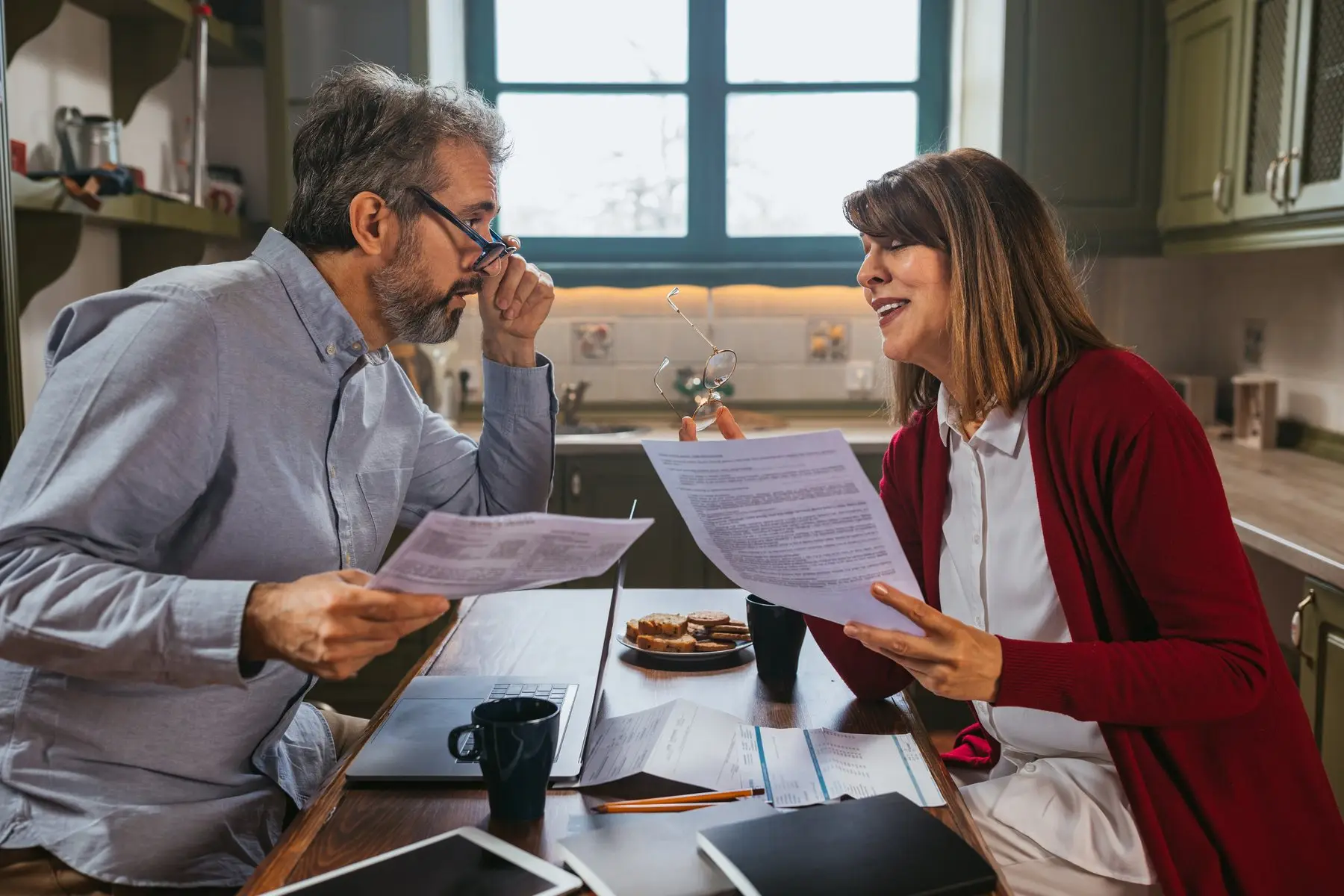 Un homme et une femme comparent des documents imprimés, assis face à face à la table d’une cuisine, un ordinateur portable entre eux