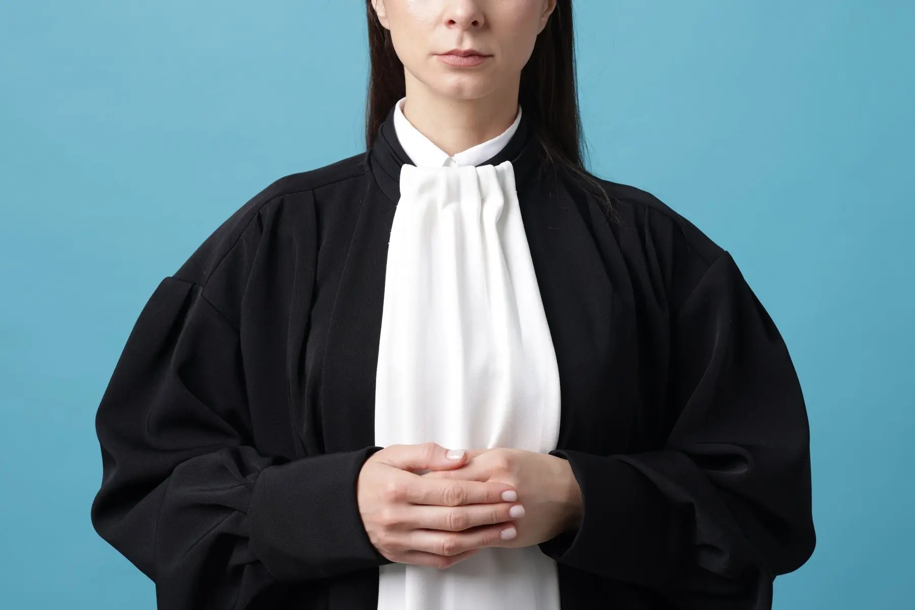 Une avocate en robe noire et rabat blanc, les mains jointes devant elle, sur un fond bleu uni