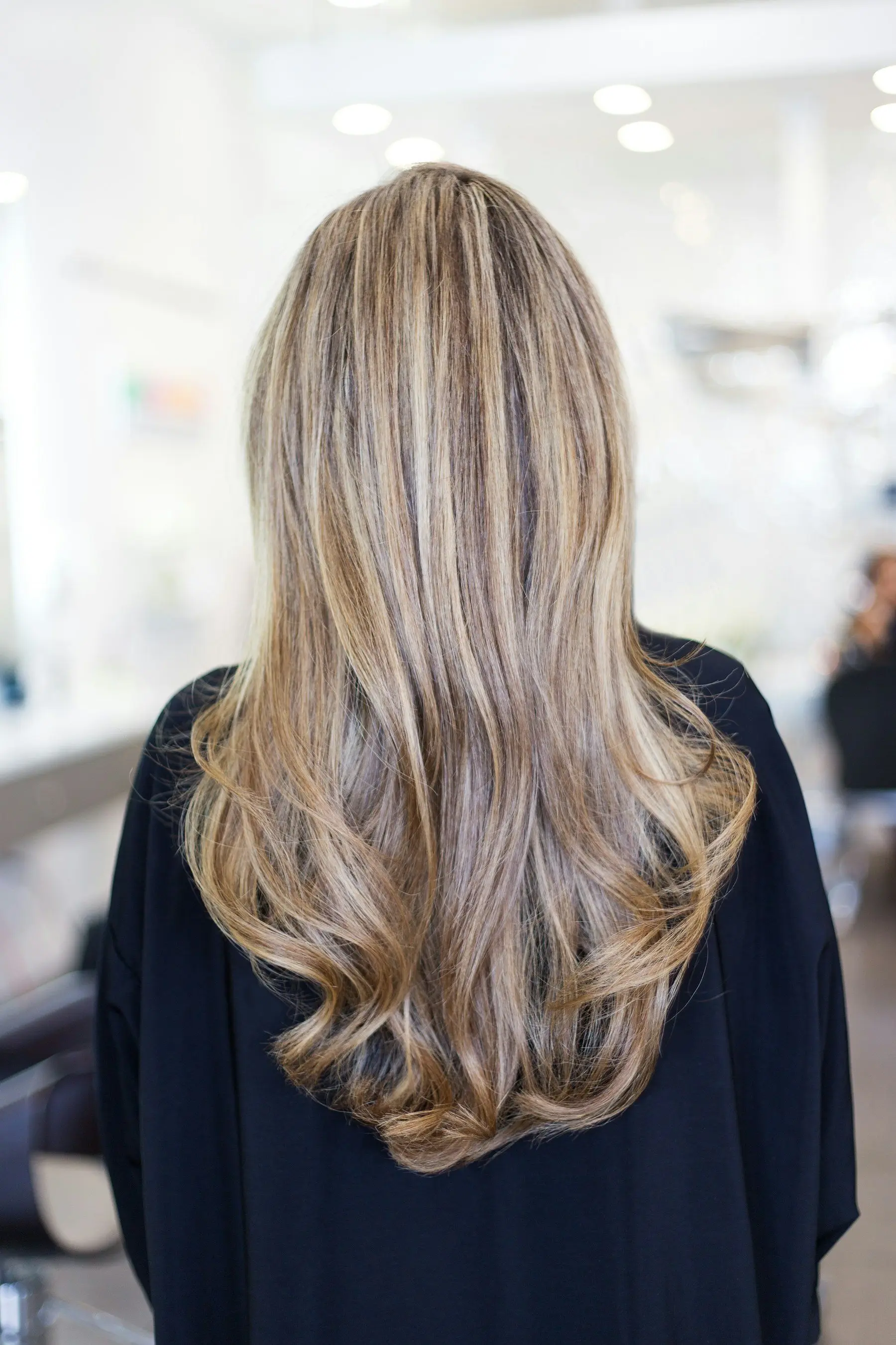 Cheveux longs blonds balayés, ondulés en pointes, vus de dos dans un salon lumineux