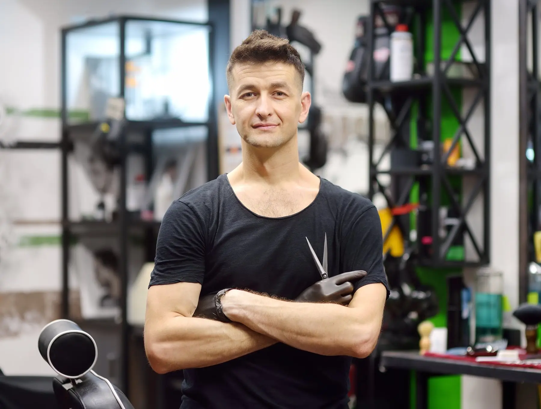 Un coiffeur en tee-shirt noir, bras croisés et ciseaux à la main, devant les étagères métalliques d’un salon