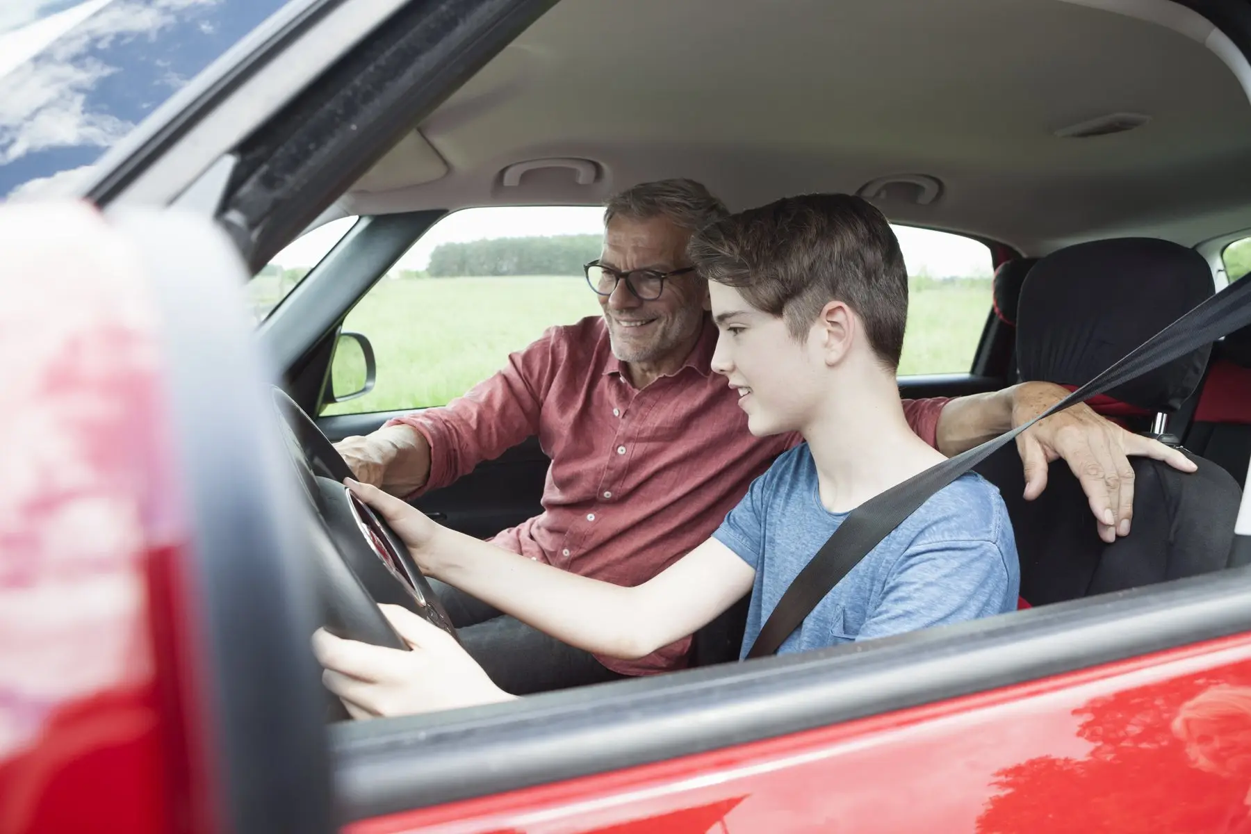 Un adolescent au volant d’une voiture rouge, à côté d’un homme à lunettes qui lui sourit, en pleine campagne