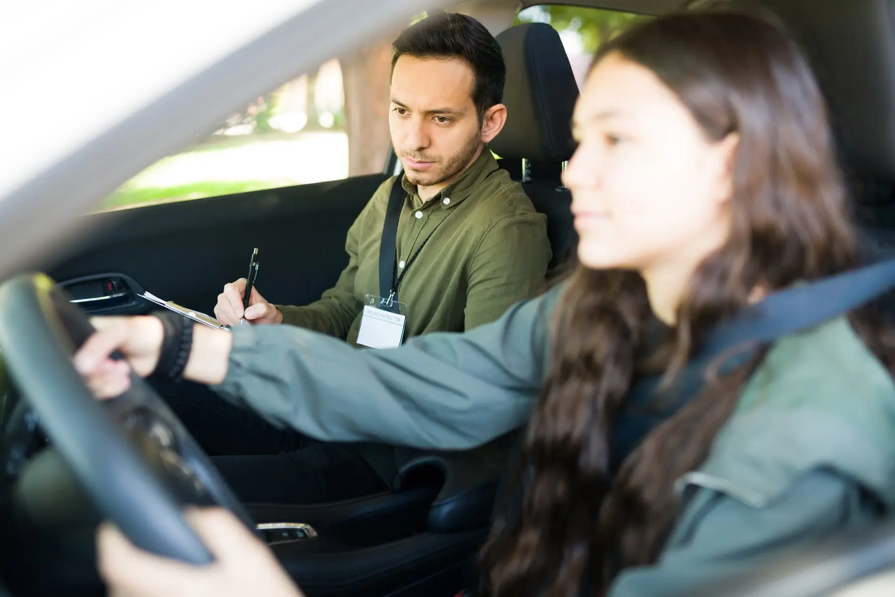 Une jeune conductrice tient le volant pendant que son moniteur, assis à côté d’elle, observe la route et note sur un bloc