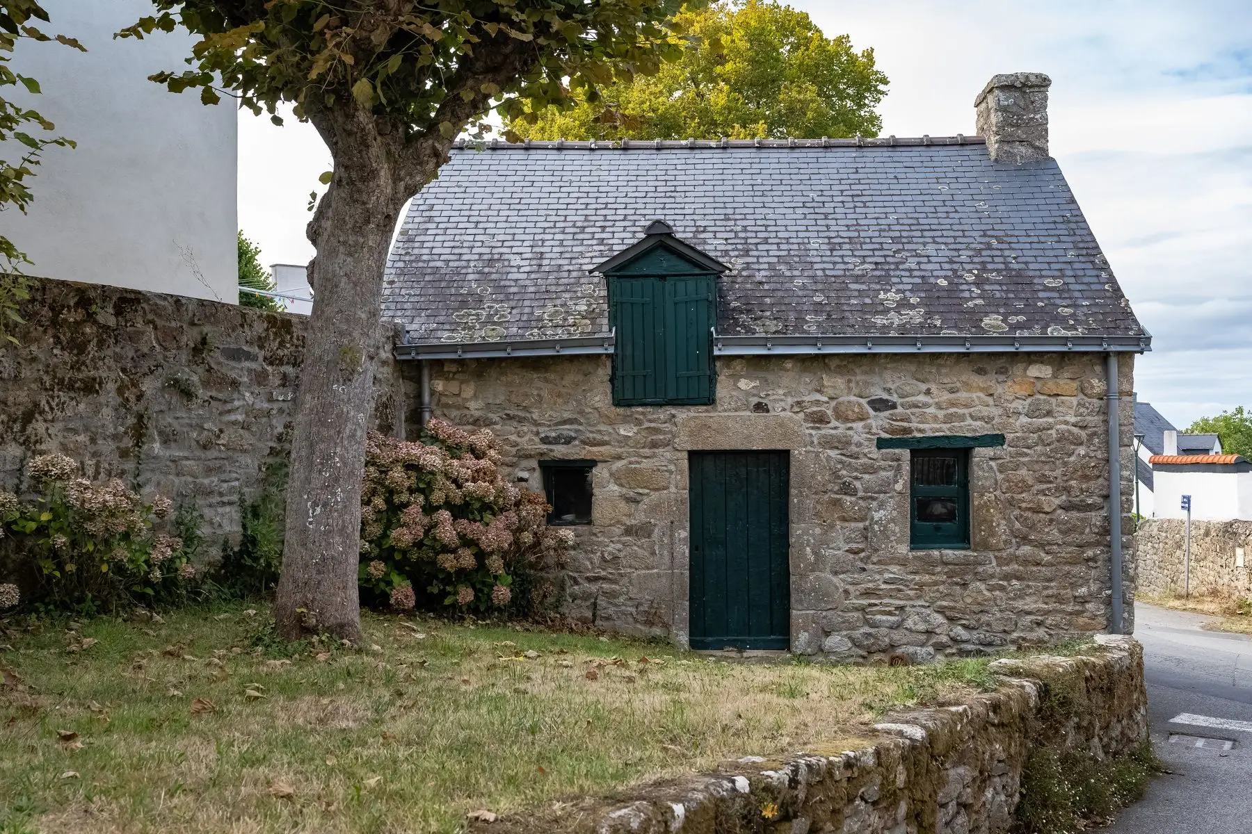 Petite maison de pierre au toit d’ardoises neuf, lucarne et porte peintes en vert, hortensias le long du mur