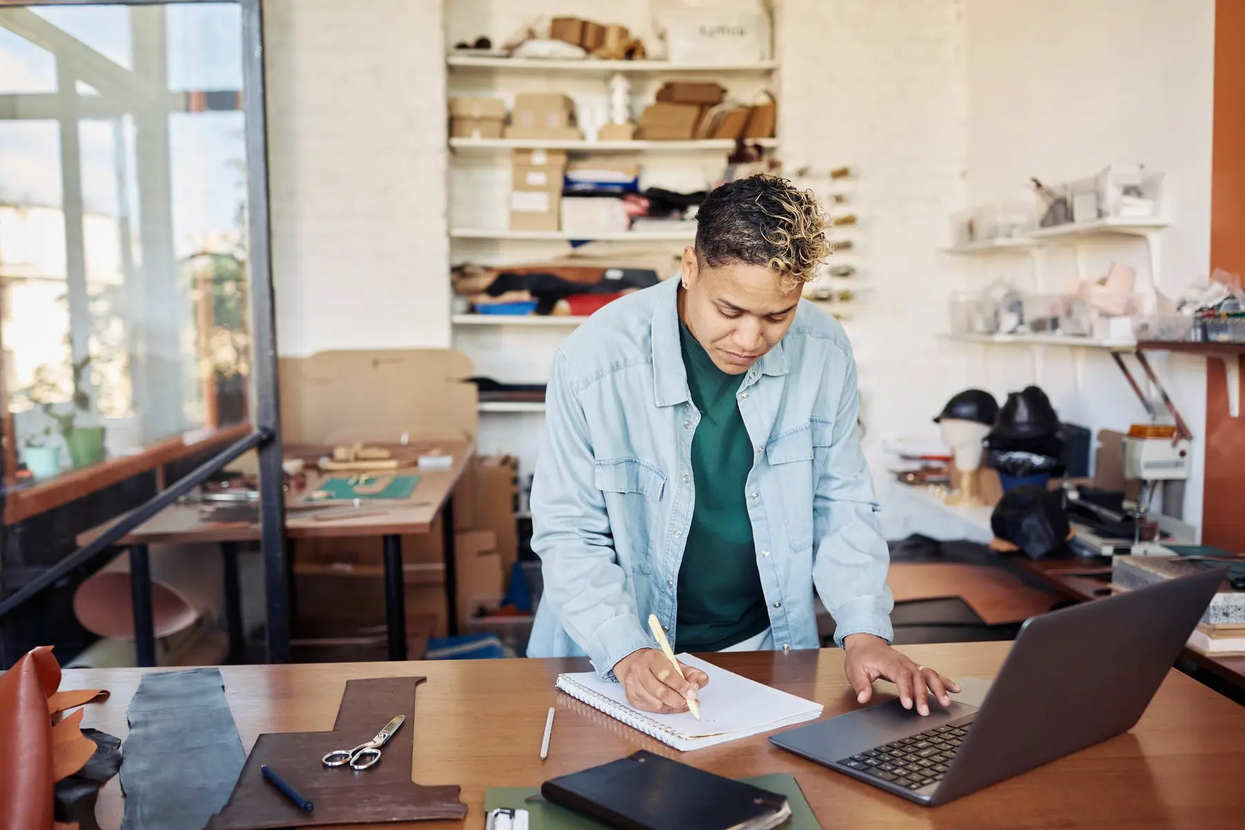 Une créatrice d’entreprise prend des notes à côté de son ordinateur portable, dans un atelier de maroquinerie aux étagères chargées