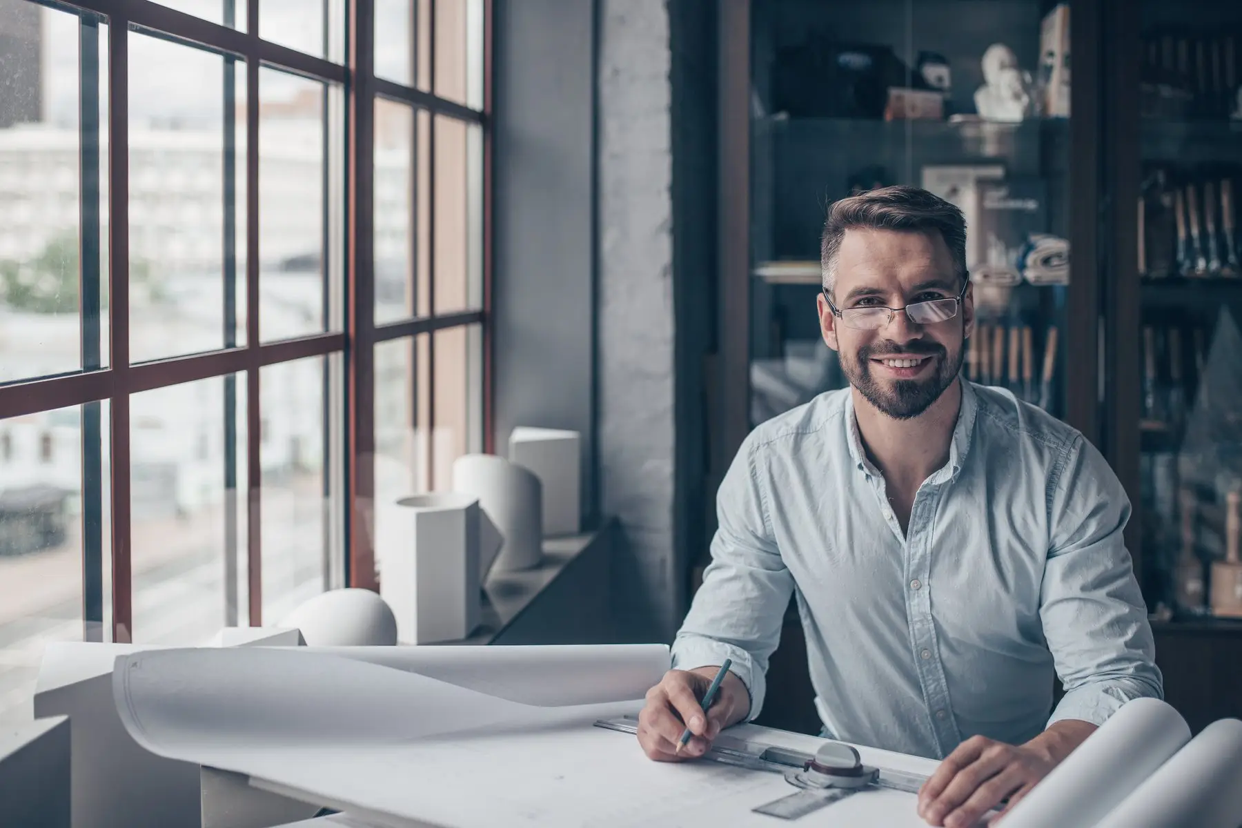 Un architecte souriant, chemise claire et lunettes, assis à sa table à dessin couverte de plans, près d’une grande fenêtre
