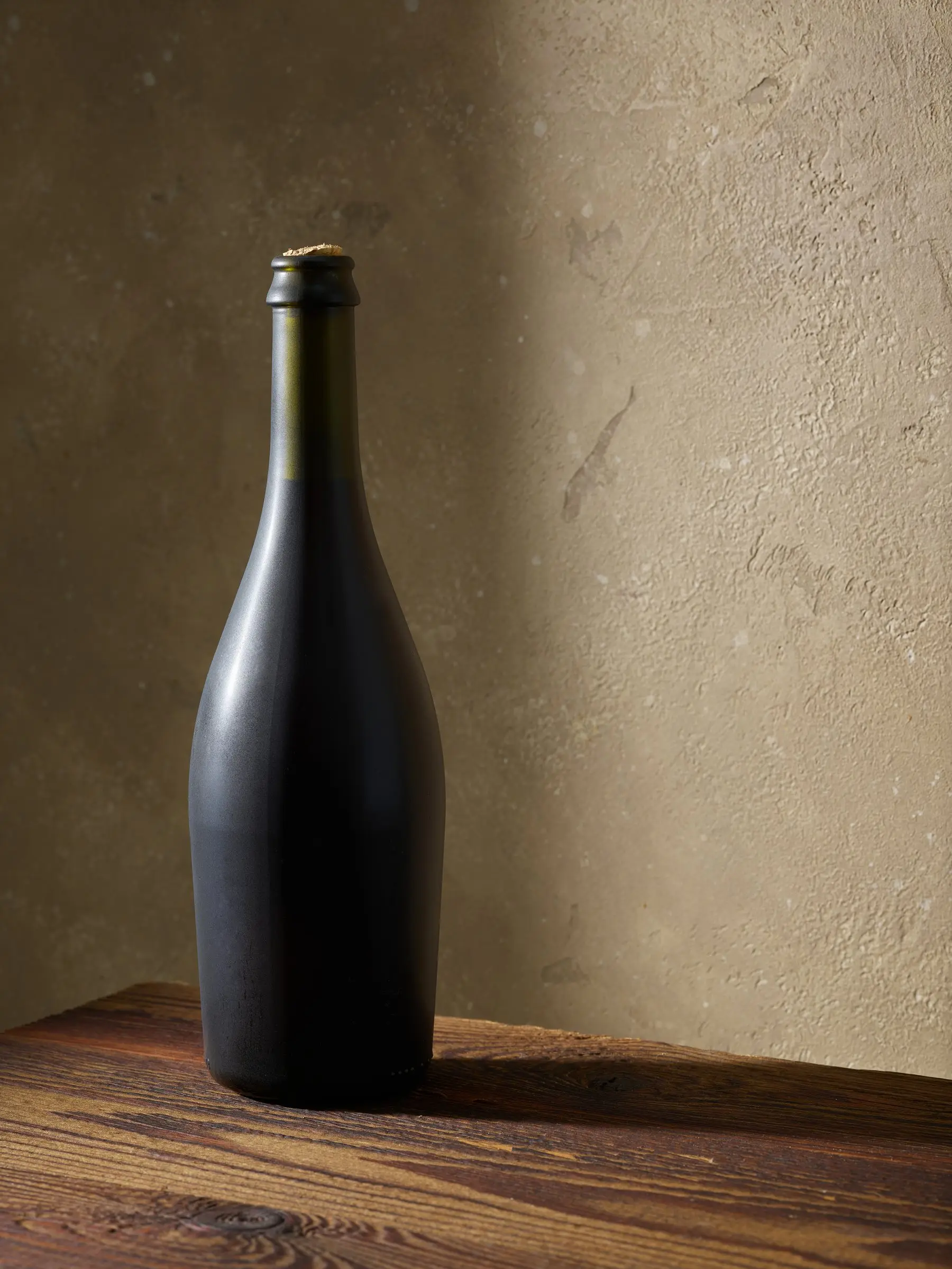 Une bouteille de verre sombre, bouchon de liège apparent, posée sur une vieille table en bois devant un mur enduit