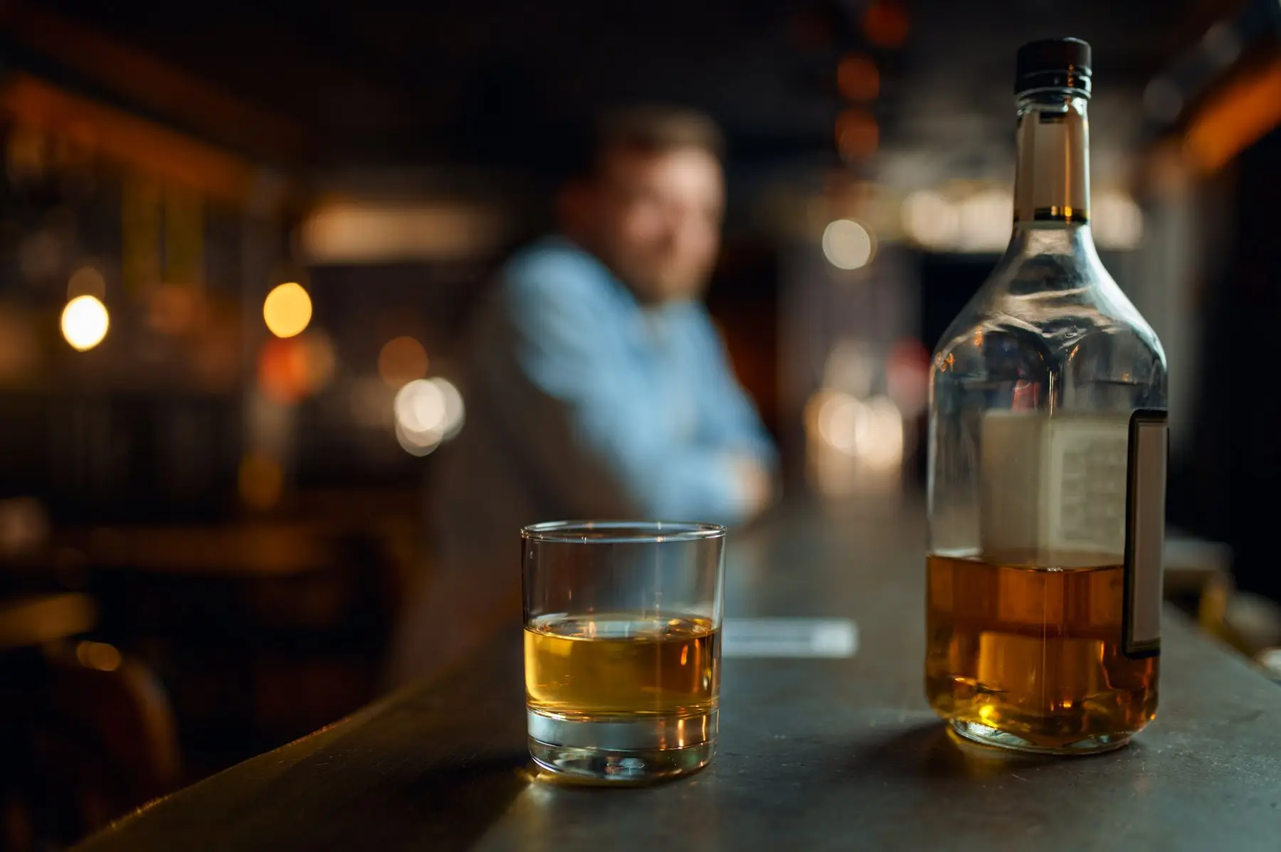 Un verre d’eau-de-vie ambrée et une bouteille à moitié pleine sur un comptoir en bois, un homme flou au fond de la salle