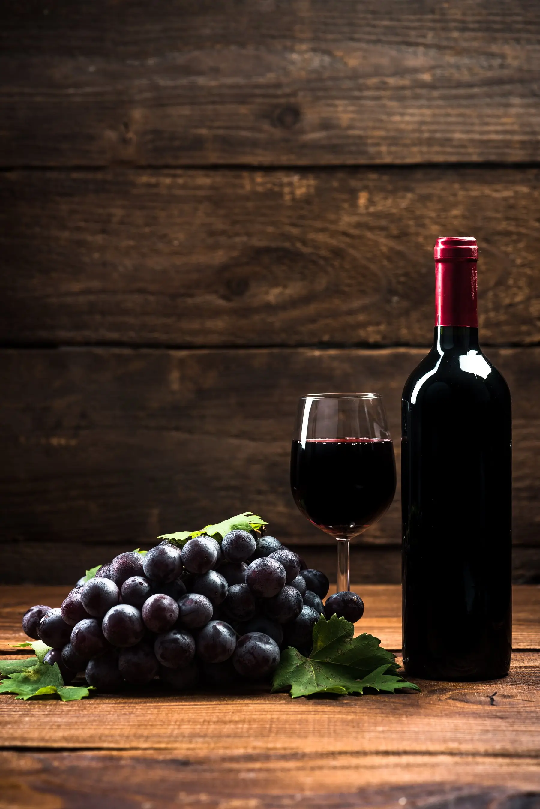 Une bouteille de vin rouge et un verre servi sur une table en bois sombre, à côté d’une grappe de raisin noir et de feuilles de vigne