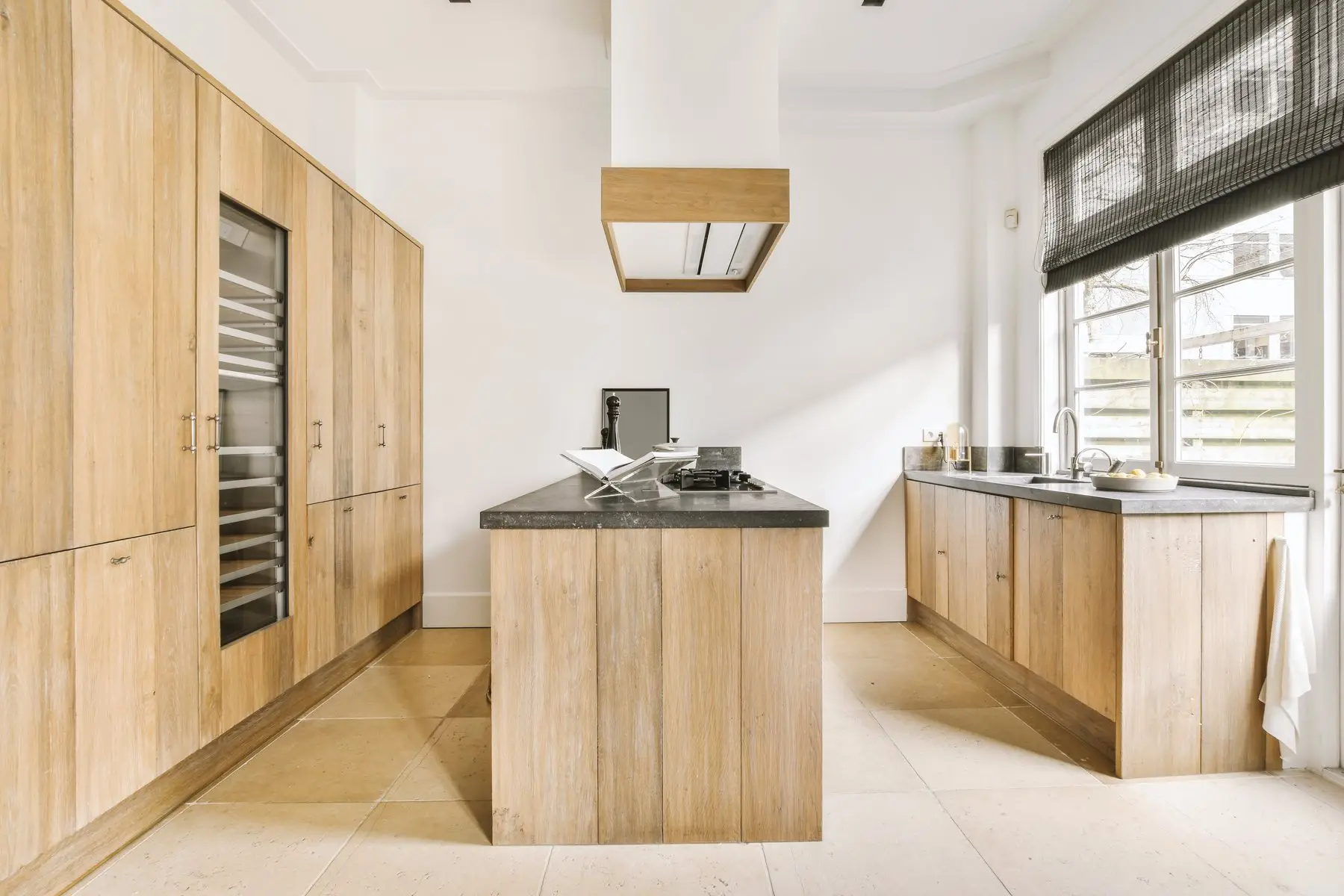 Une cuisine aux grandes façades en chêne massif, un îlot en planches de chêne avec plaque de cuisson et plan de travail sombre, une grande fenêtre à store sombre