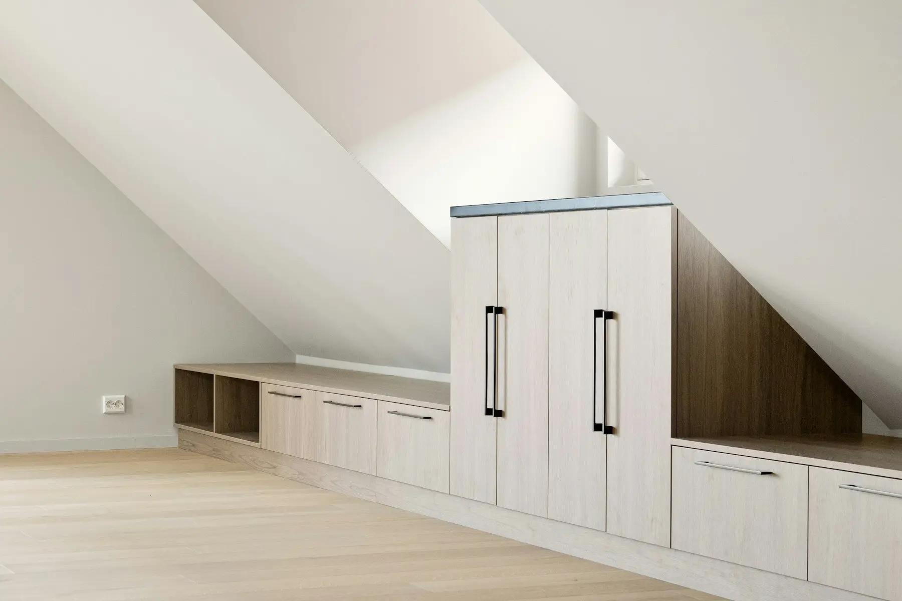 Des rangements sous une pente de toit : penderie à deux portes, tiroirs bas et niches ouvertes en bois clair, poignées noires