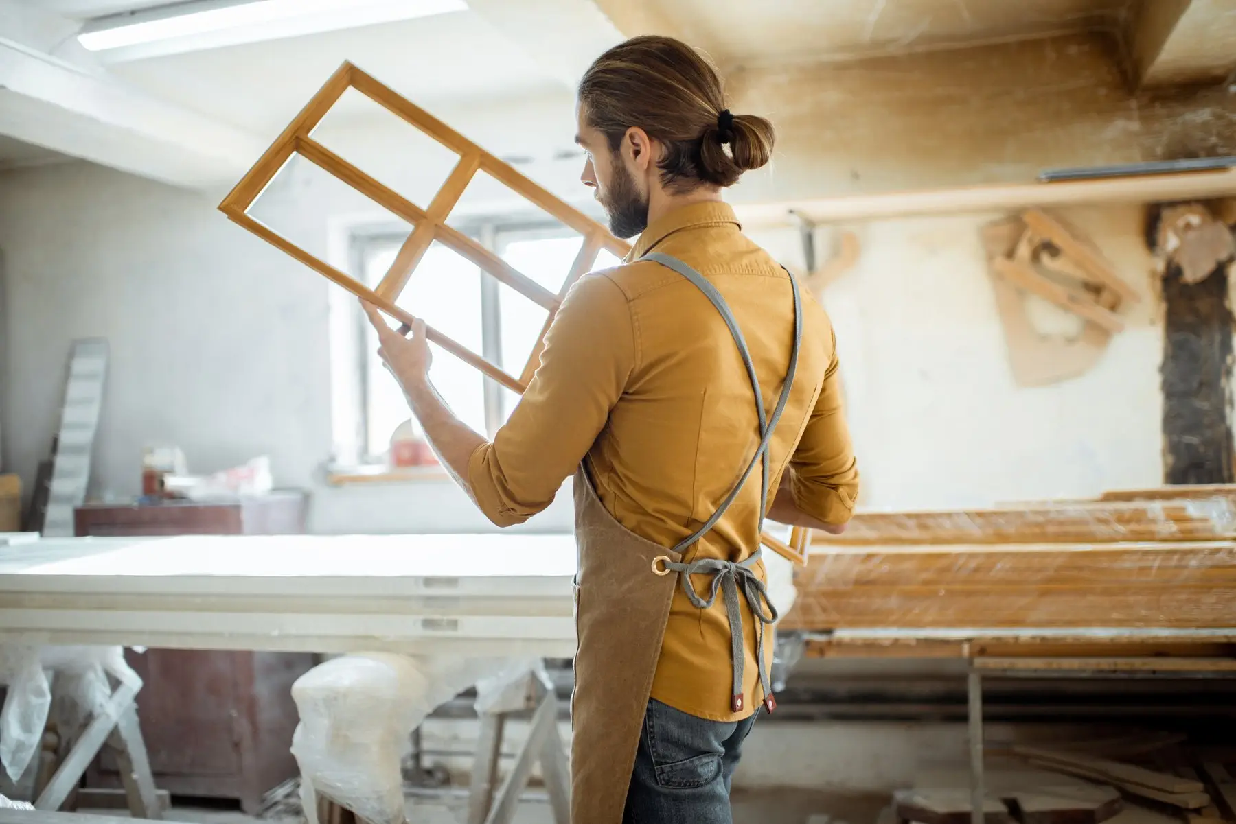 Dans l’atelier, un menuisier en tablier tient à bout de bras un châssis de fenêtre à petits bois en bois clair