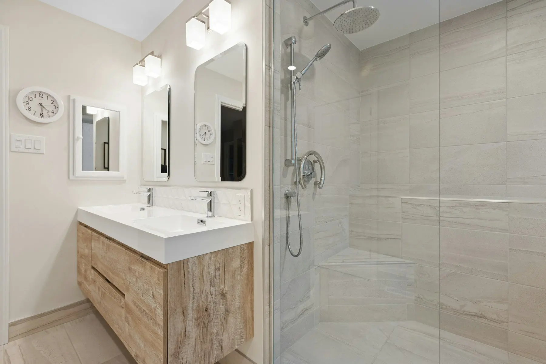 Une salle de bain claire : meuble vasque en bois, deux miroirs, horloge murale et grande douche derrière une paroi vitrée