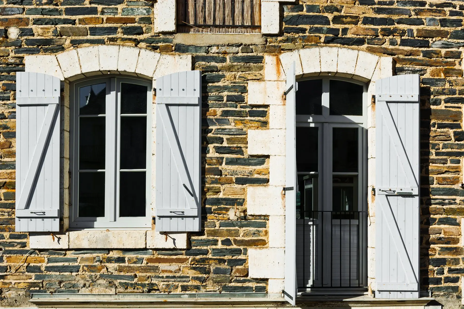 Façade de pierre sombre aux encadrements clairs, fenêtres à petits carreaux et volets gris perle ouverts