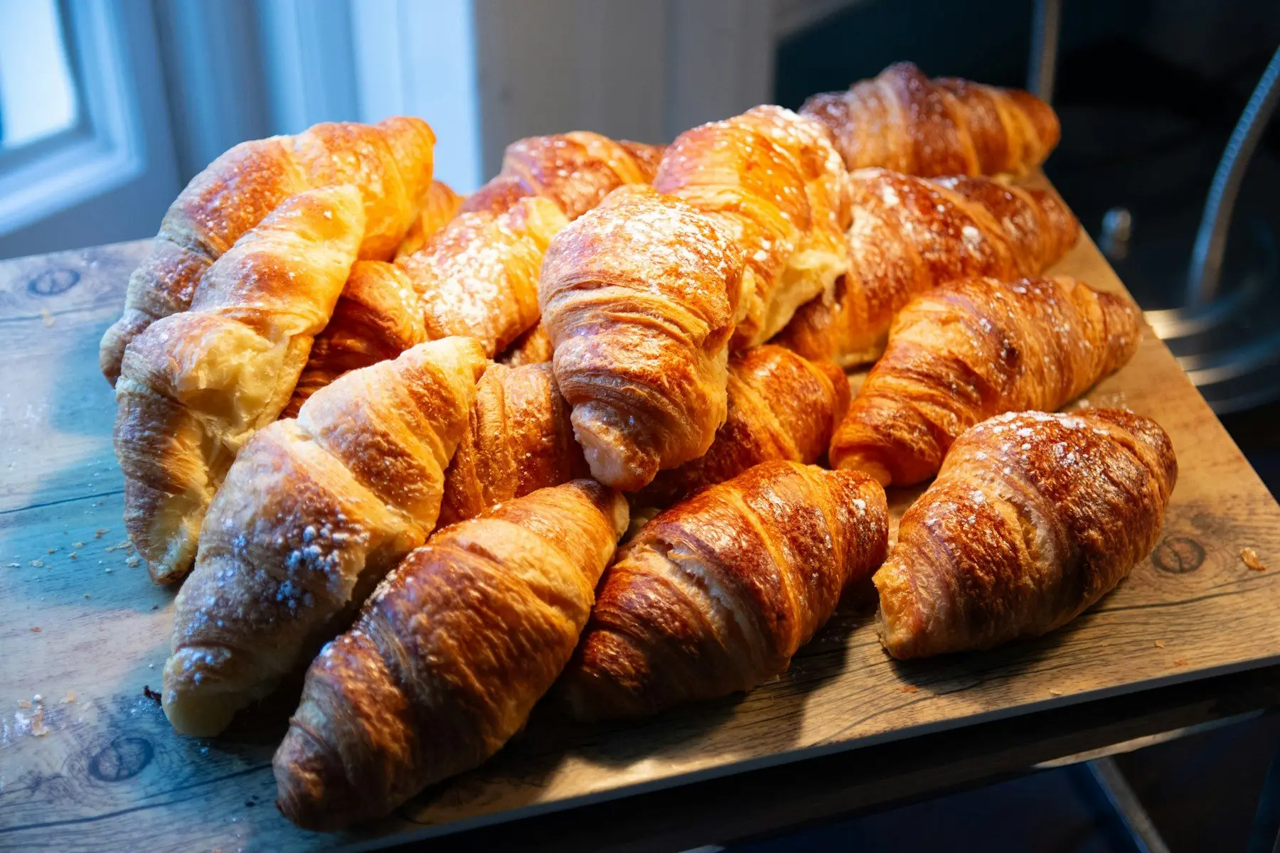 Croissants dorés saupoudrés de sucre glace sur une planche en bois, près d’une fenêtre