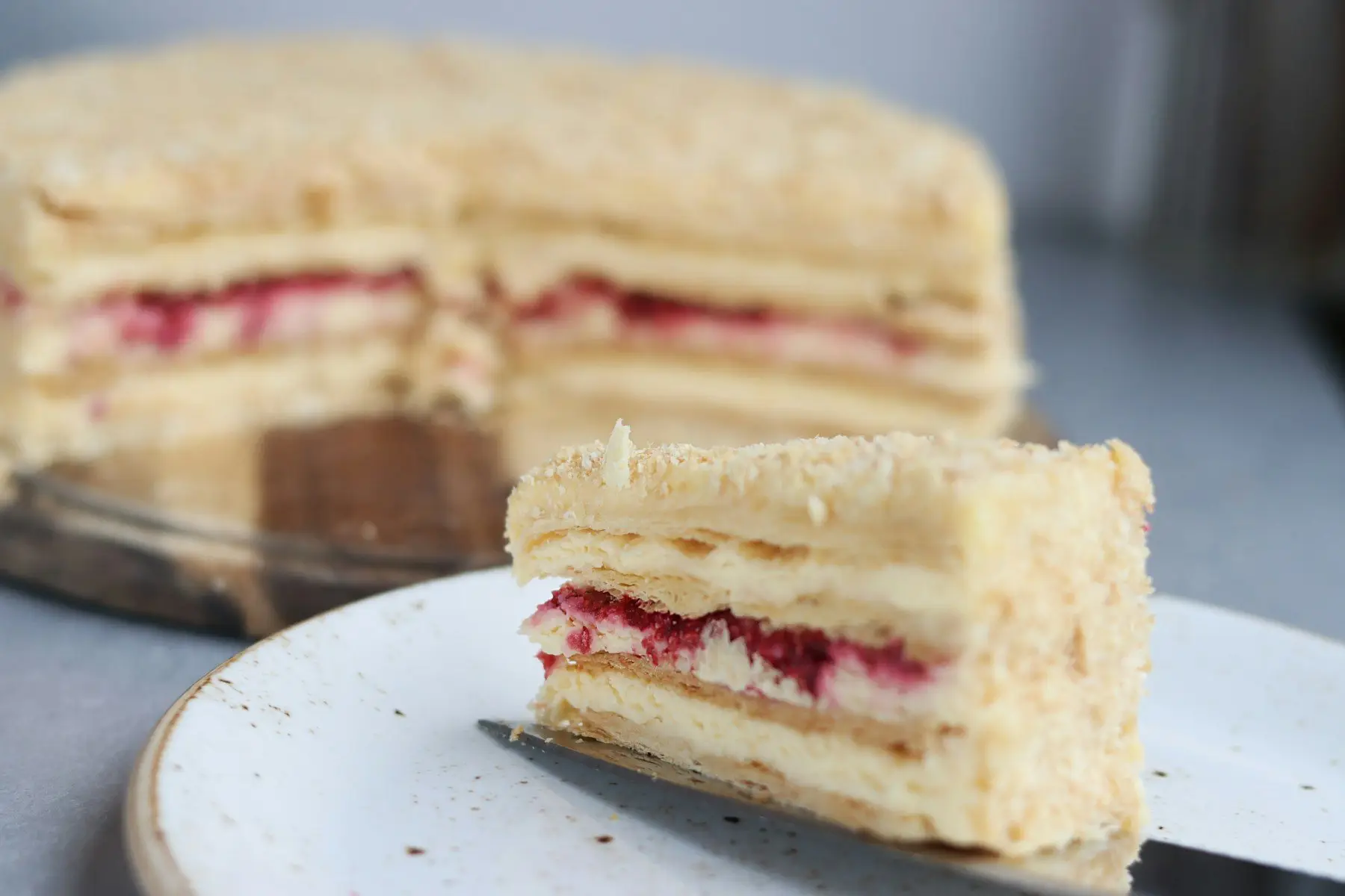 Une part de gâteau feuilleté à la crème et aux framboises sur une assiette mouchetée, le reste du gâteau en arrière-plan