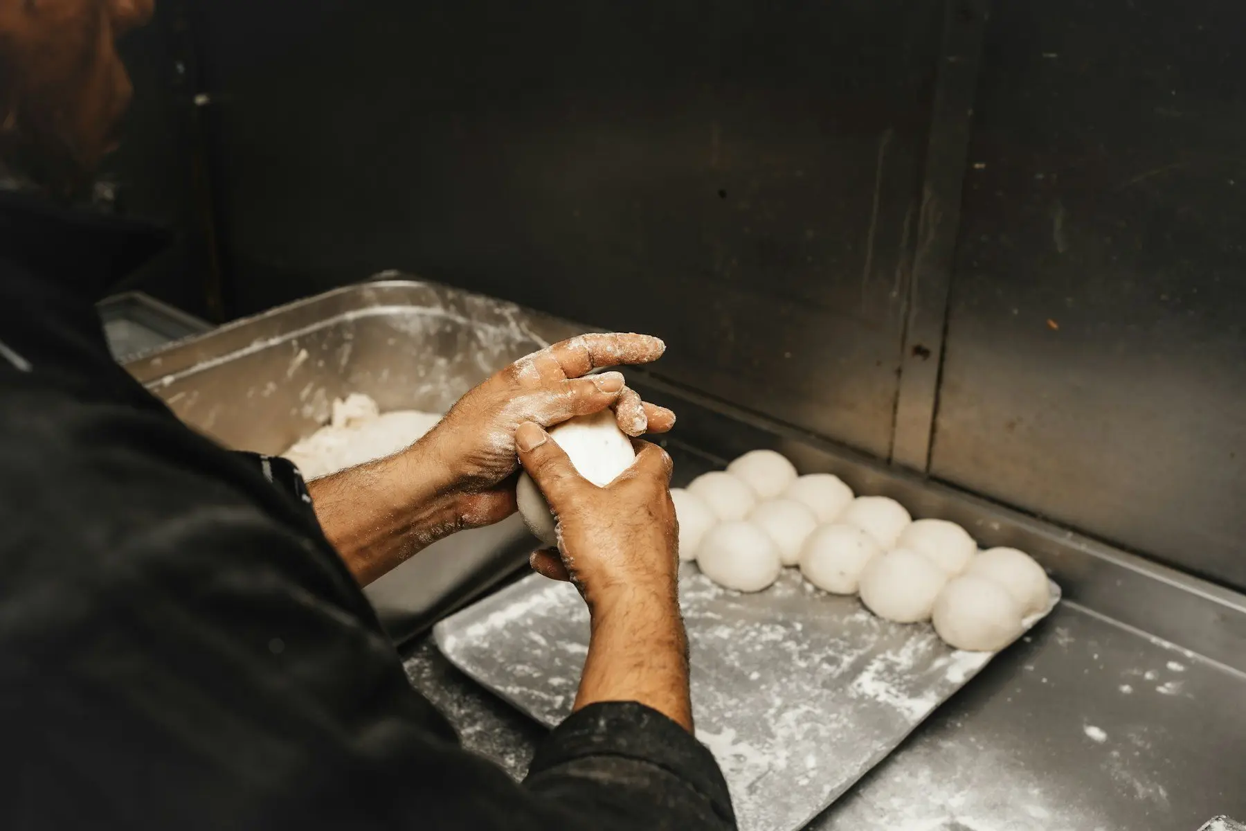 Les mains farinées d’un boulanger boulent un pâton à côté d’une plaque de petits pâtons bien ronds