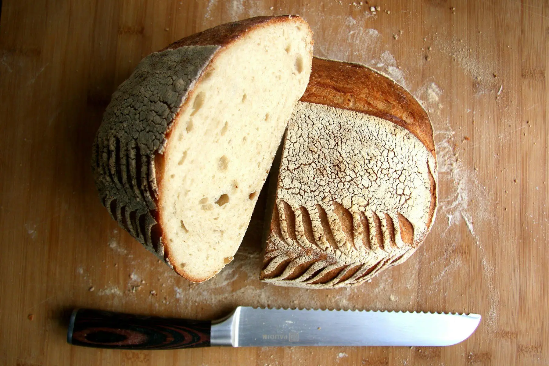 Une miche de campagne coupée en deux sur une planche farinée, mie alvéolée et croûte grignée, couteau à pain posé devant