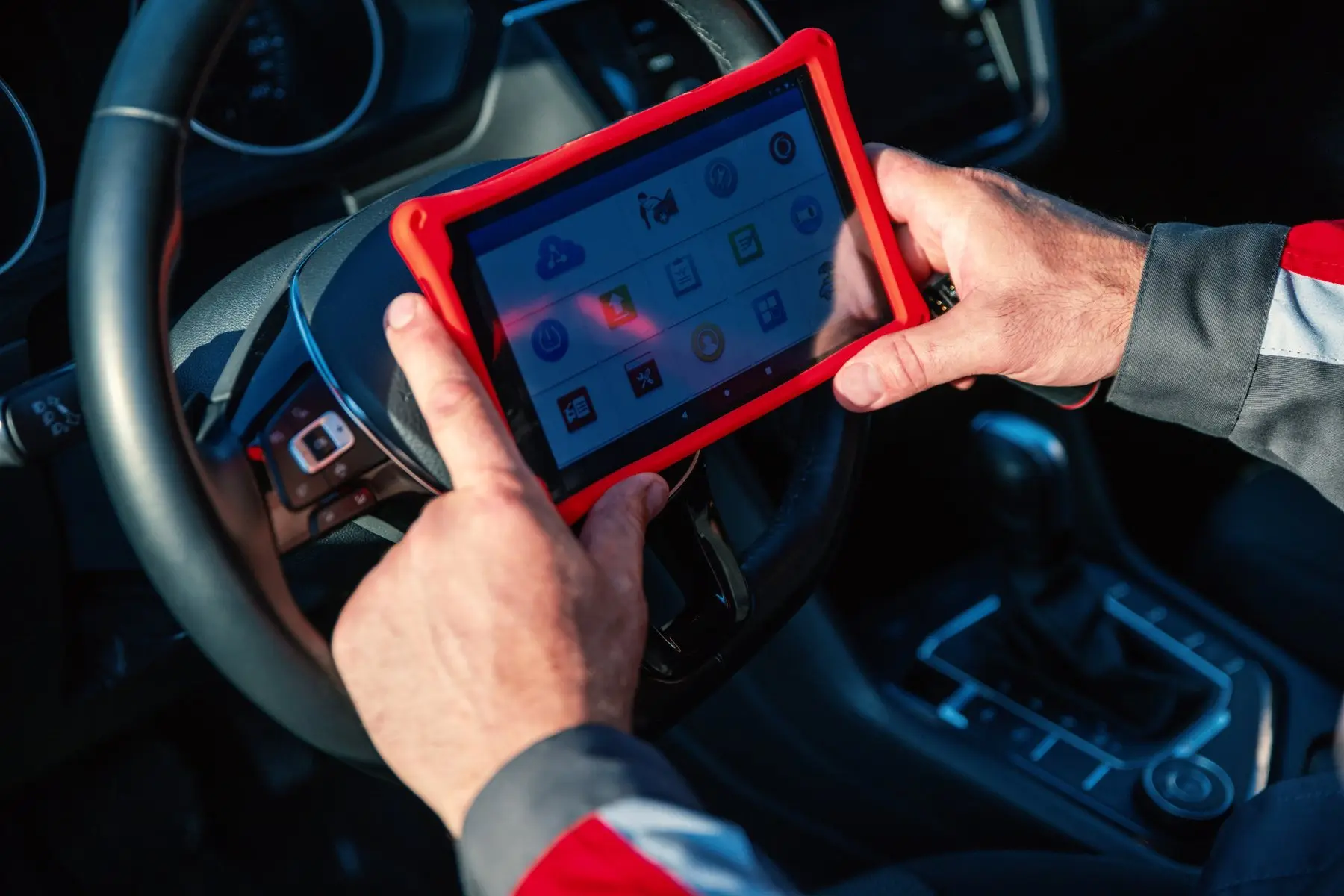 Des mains tiennent une tablette de diagnostic à coque rouge devant le volant d’une voiture