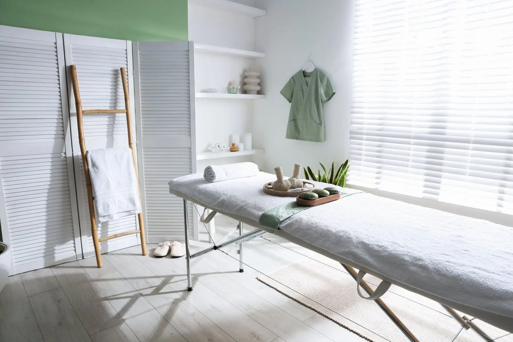 Cabine de soin lumineuse : table recouverte de serviettes blanches, galets et pochons aux herbes, paravent blanc et mur vert sauge