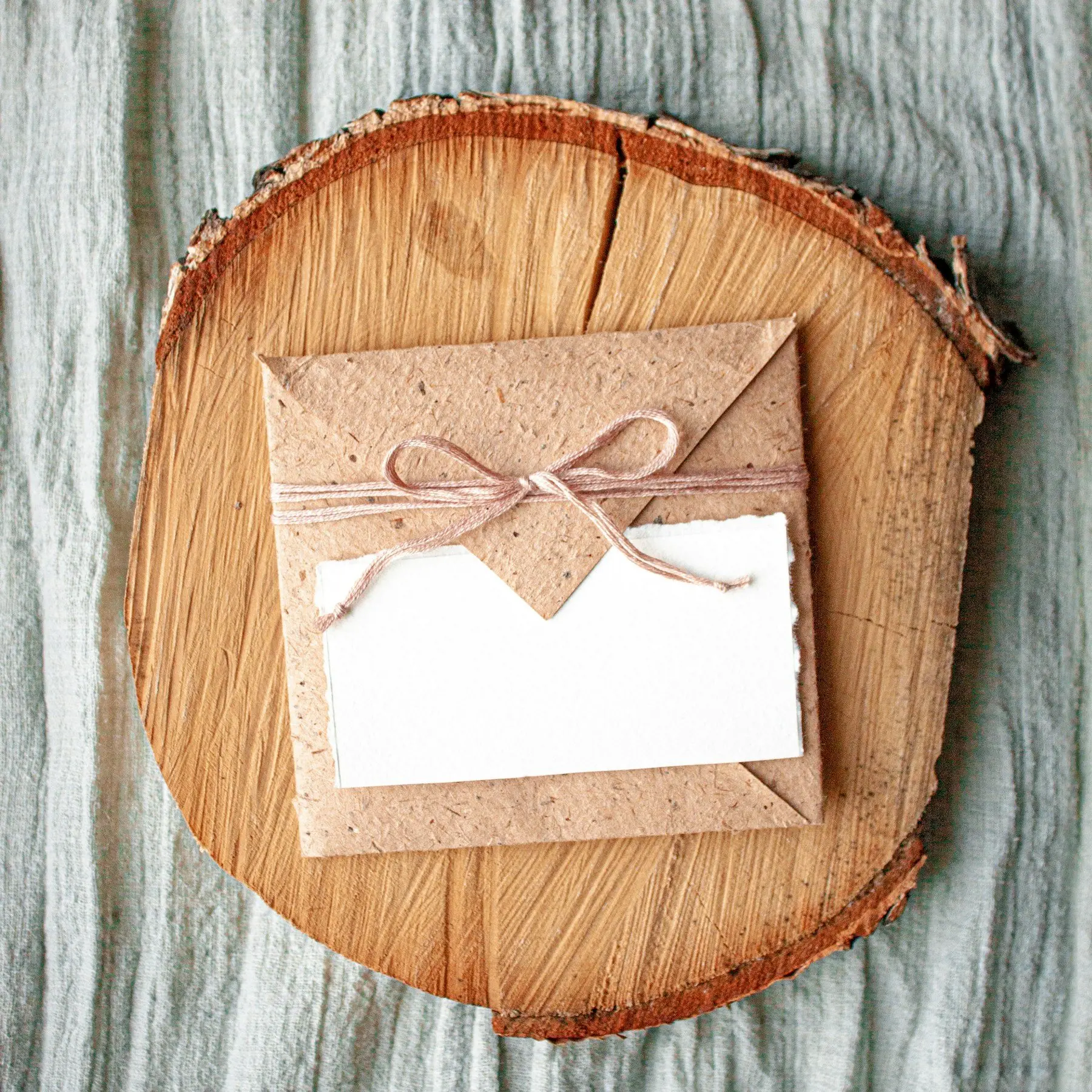 Enveloppe en papier kraft nouée d’une ficelle rose et carte blanche, posées sur une rondelle de bois et un lin vert sauge
