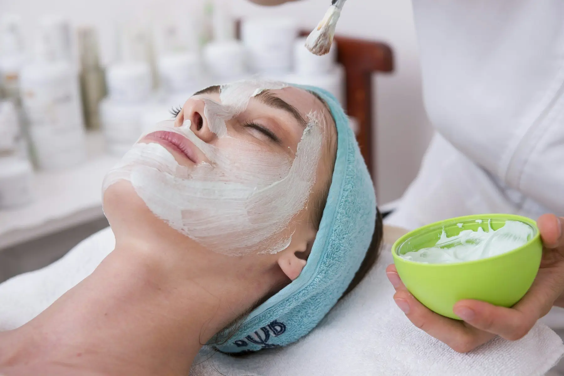 Une esthéticienne applique au pinceau un masque crème sur le visage d’une cliente allongée, les cheveux retenus par un bandeau