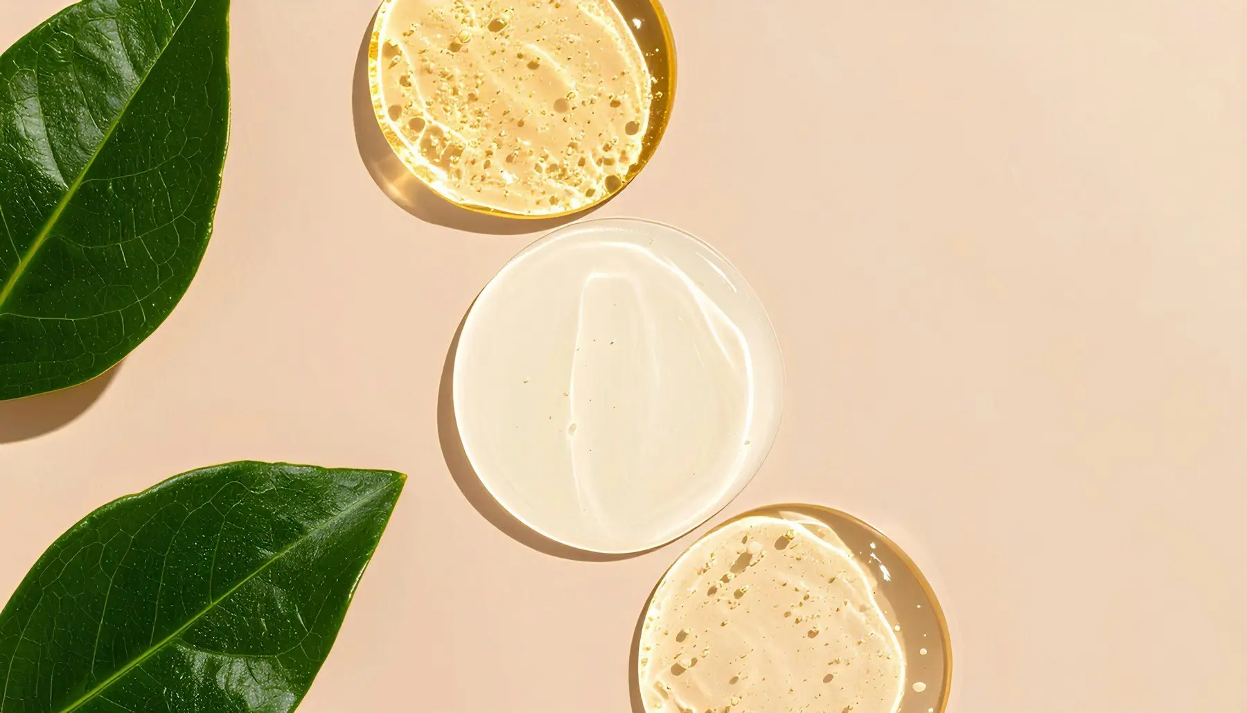 Trois touches de gel et de sérum transparents sur fond rose pâle, à côté de deux feuilles vertes