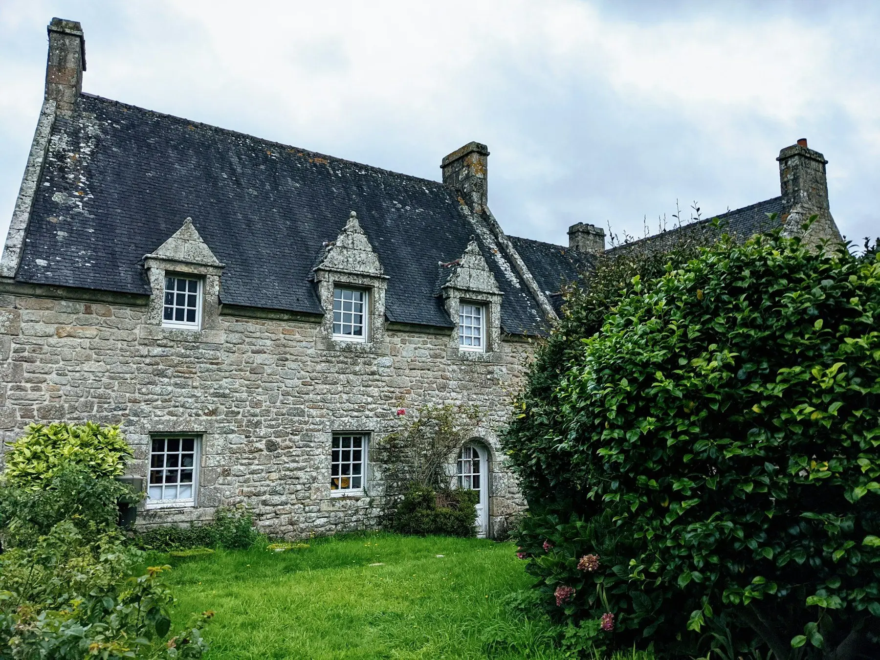 Longère en granit aux lucarnes de pierre, bordée de haies et d’arbustes, pelouse au premier plan