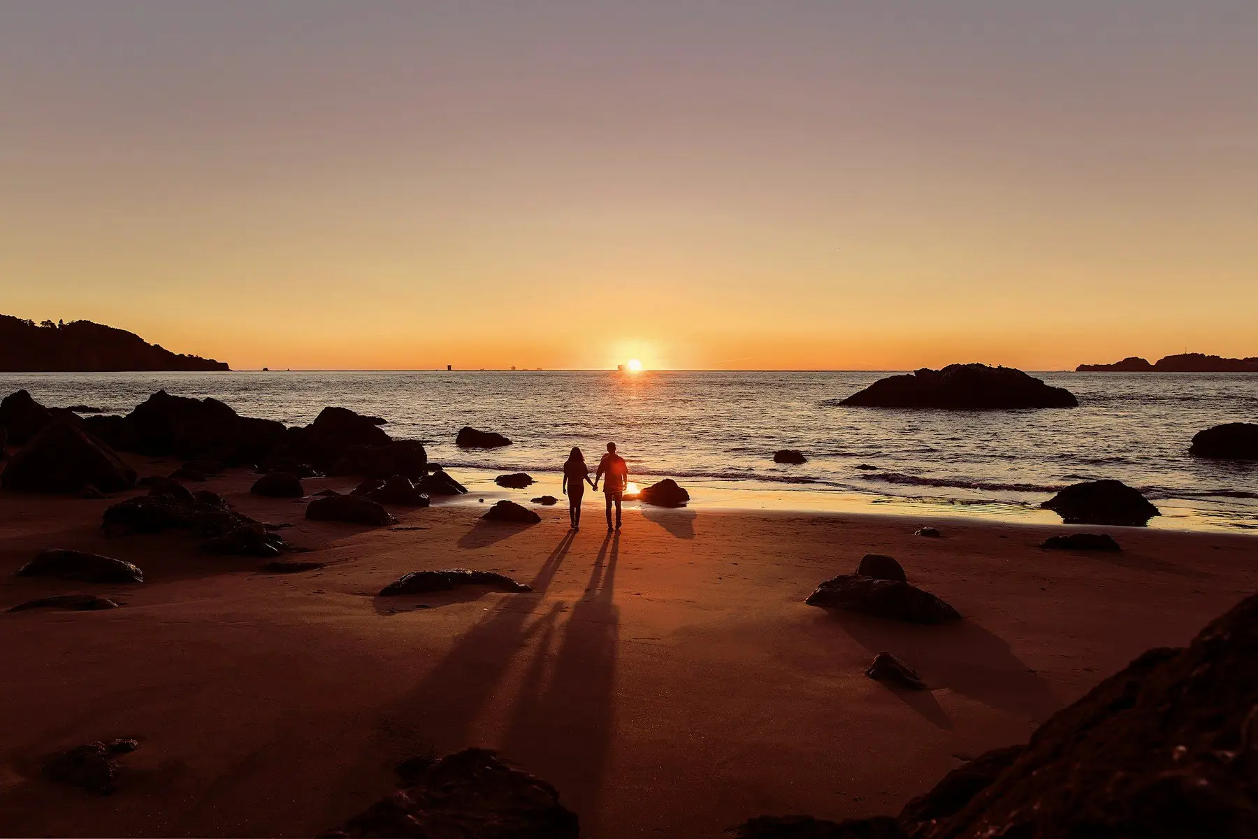 Deux silhouettes main dans la main sur une plage semée de rochers, face au soleil couchant