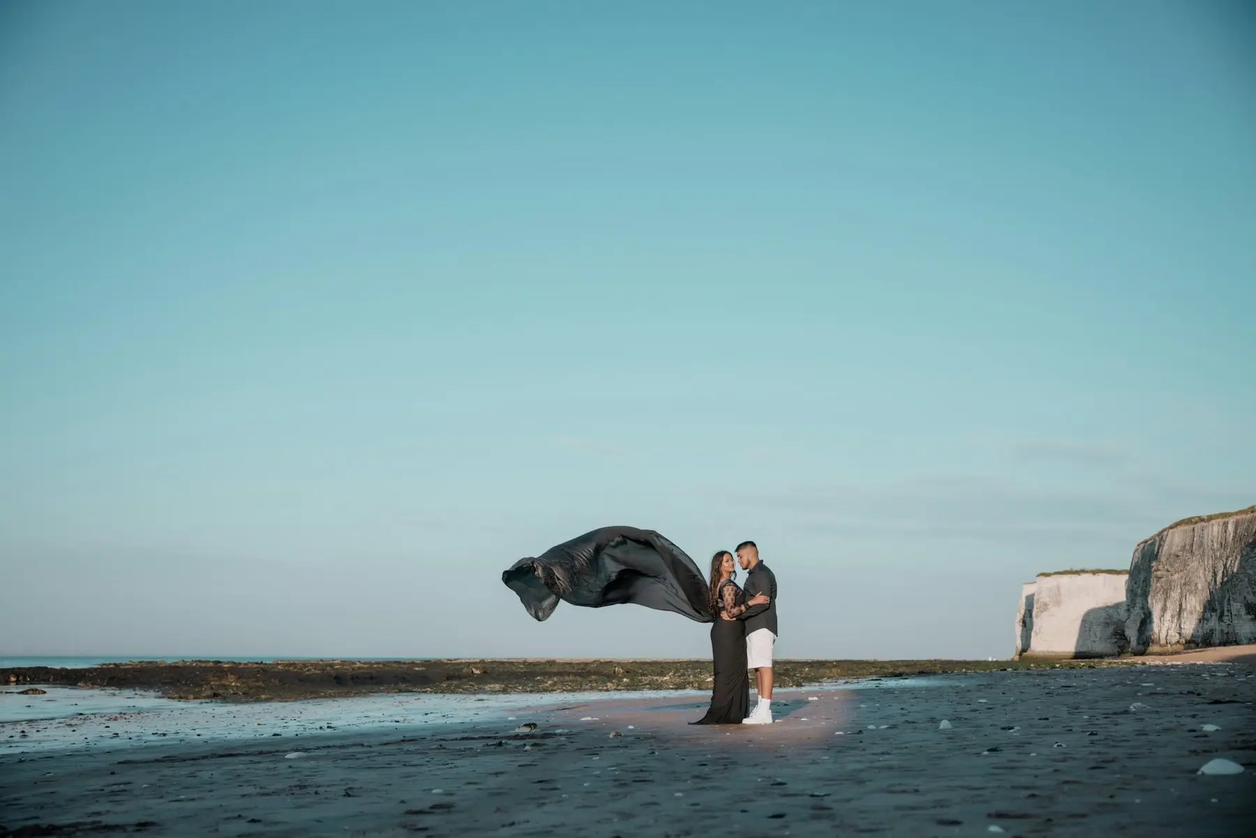 Un couple enlacé sur une plage à marée basse, le long voile sombre de la robe soulevé par le vent, des falaises blanches au loin