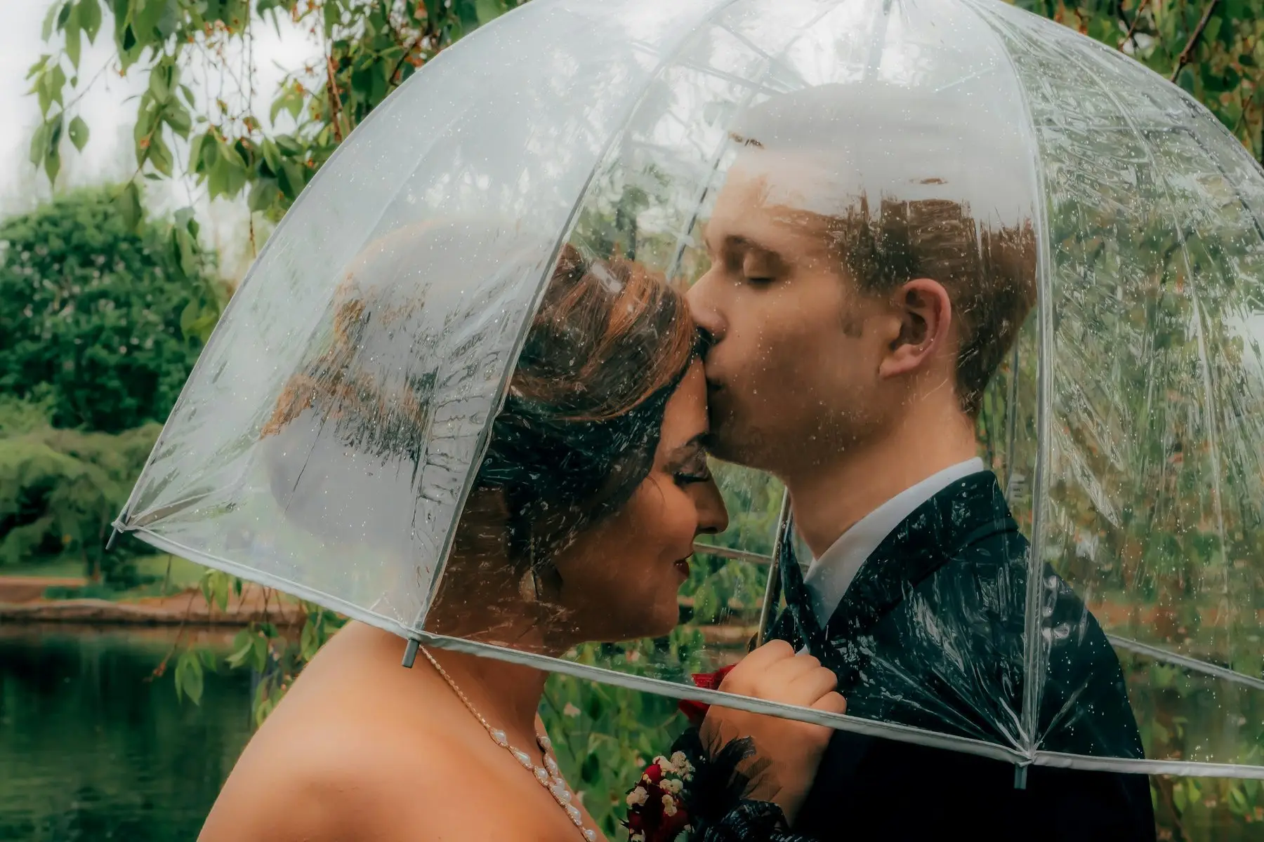 Sous un parapluie transparent couvert de gouttes, un marié embrasse le front de la mariée, devant un étang bordé d’arbres