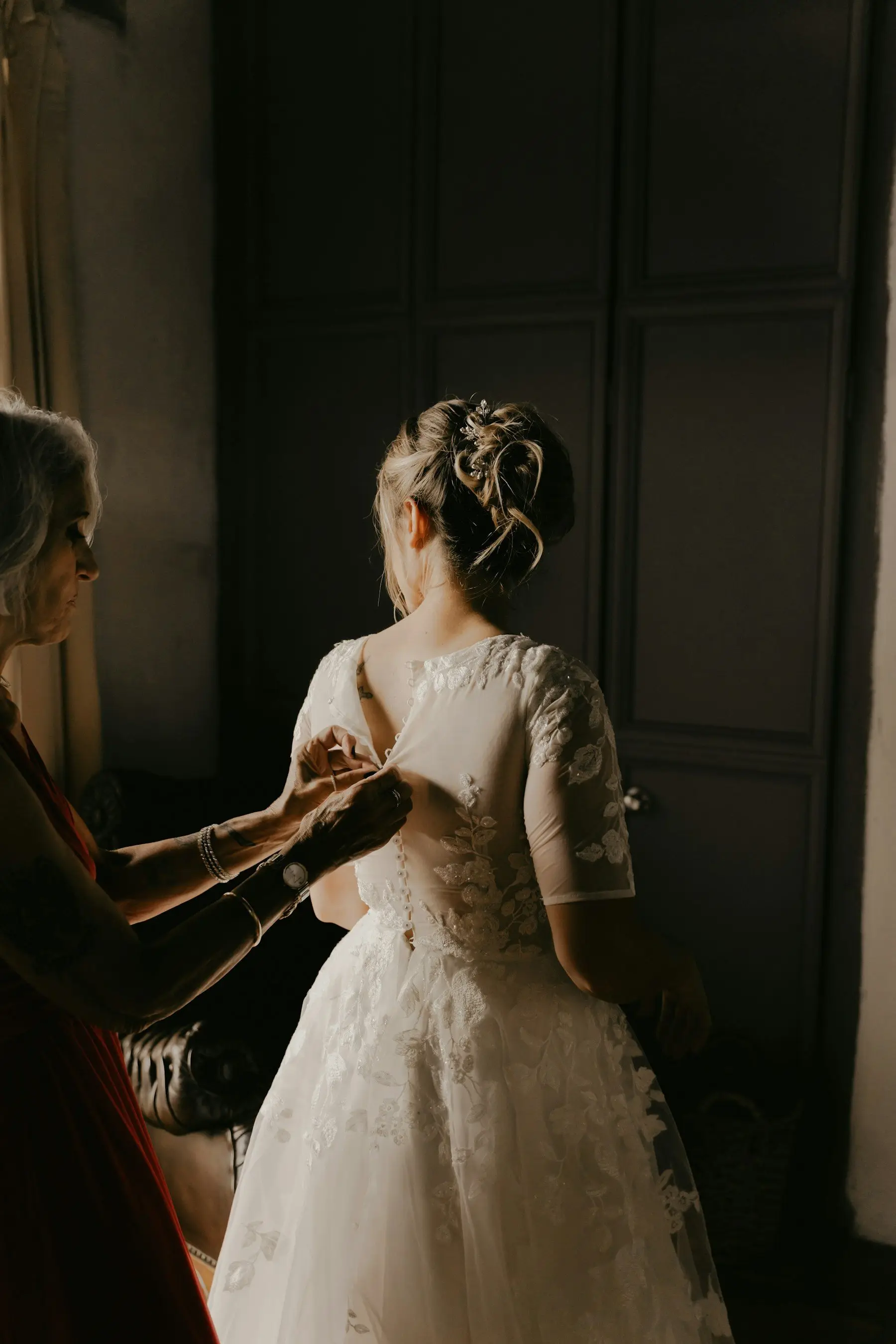 Une femme boutonne le dos de la robe en dentelle d’une mariée, près d’une fenêtre, dans une chambre plongée dans la pénombre