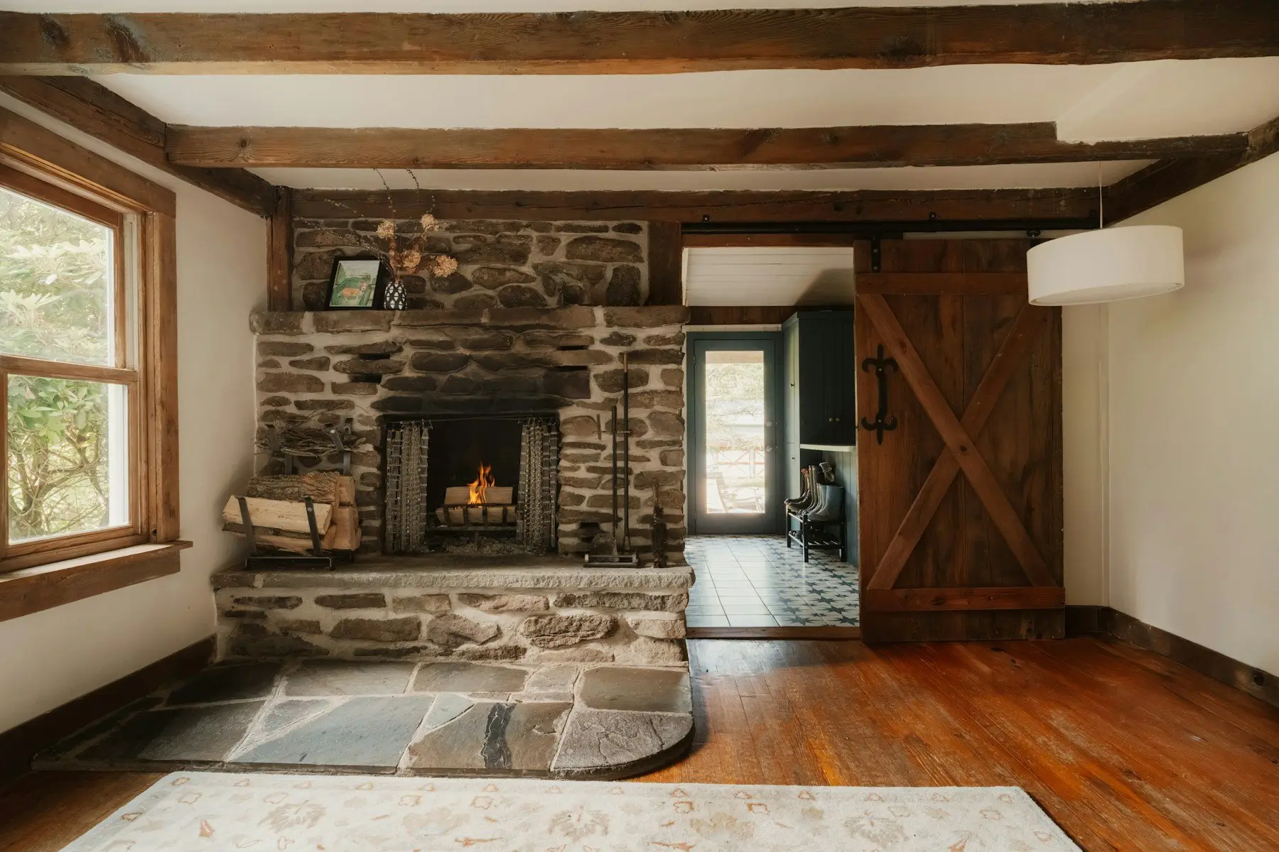 Salon sous poutres anciennes, grande cheminée en pierre où brûle un feu, porte coulissante en bois et carreaux de ciment dans l’entrée