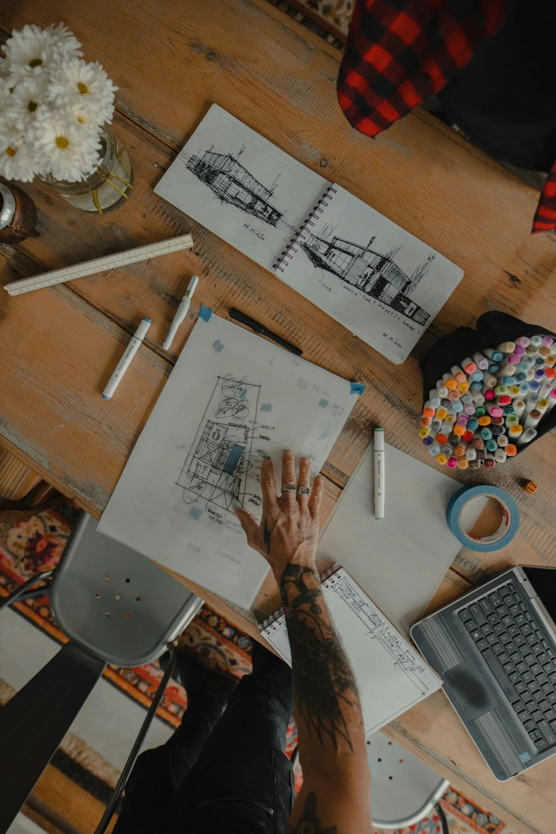Vue de dessus d’une table de travail : plan d’aménagement sur calque, carnet de croquis d’une maison, feutres et règle