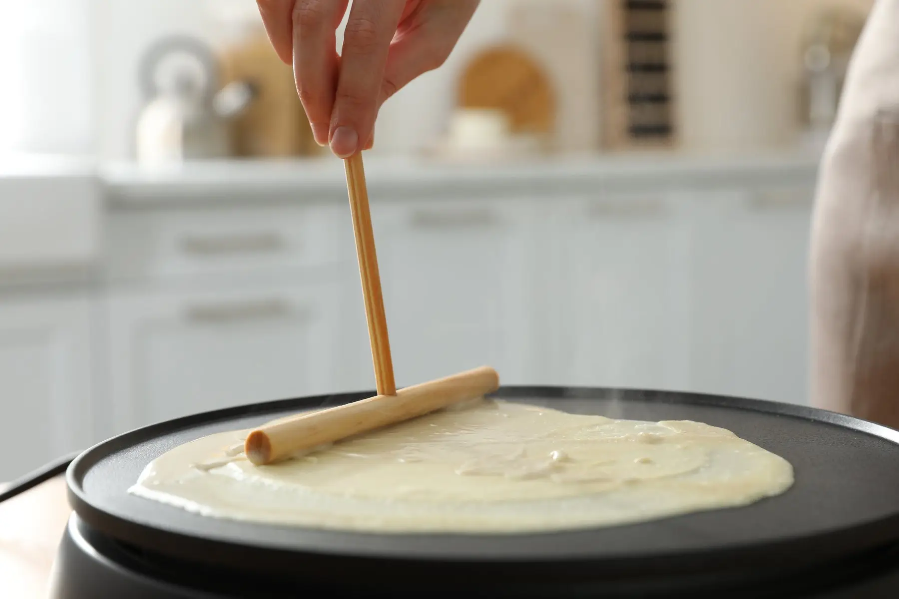 Une main étale la pâte à crêpe sur un billig avec un rozell en bois