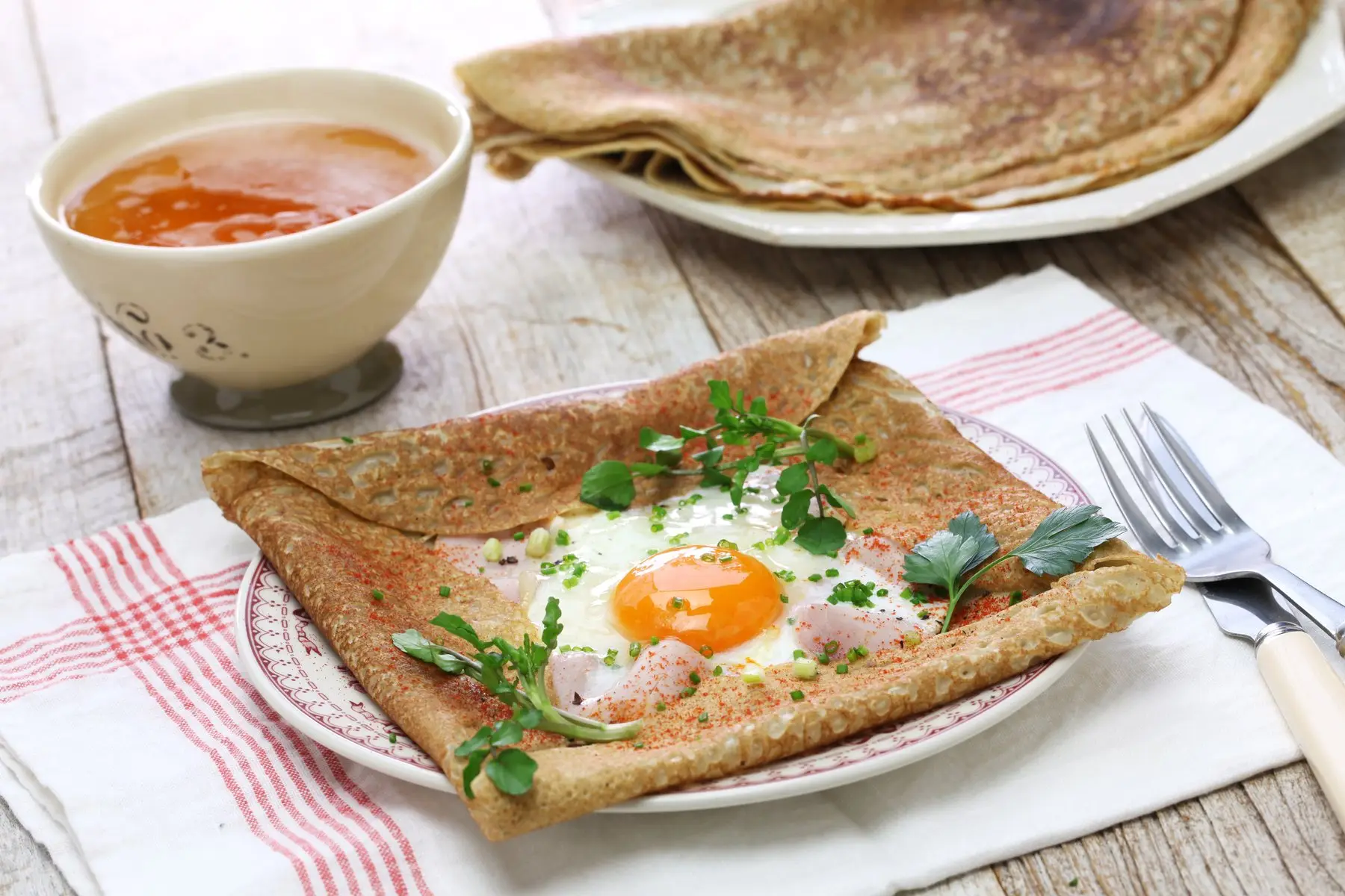 Galette de blé noir garnie d’un œuf au plat, de jambon et d’herbes fraîches, à côté d’une bolée de cidre et d’une pile de galettes