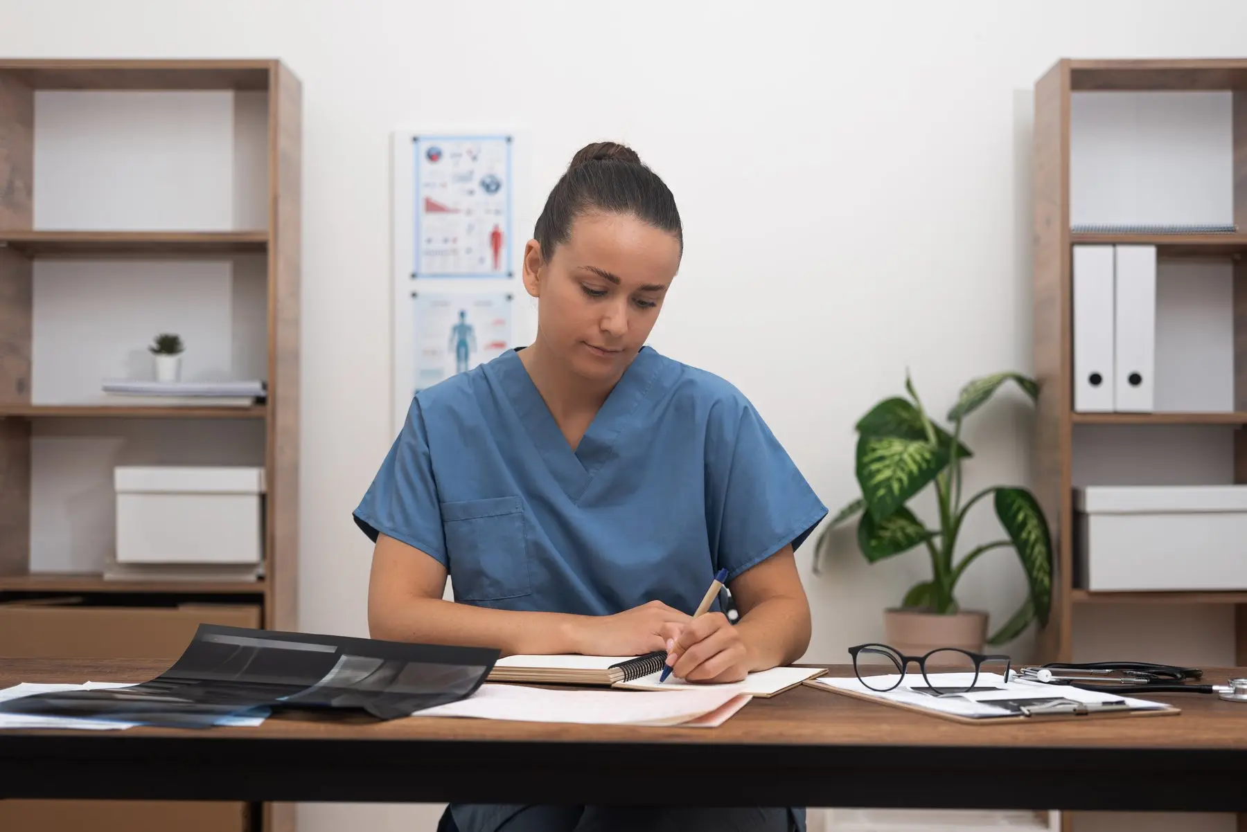 Une praticienne en tunique bleue prend des notes à son bureau, des clichés de radiographie posés devant elle
