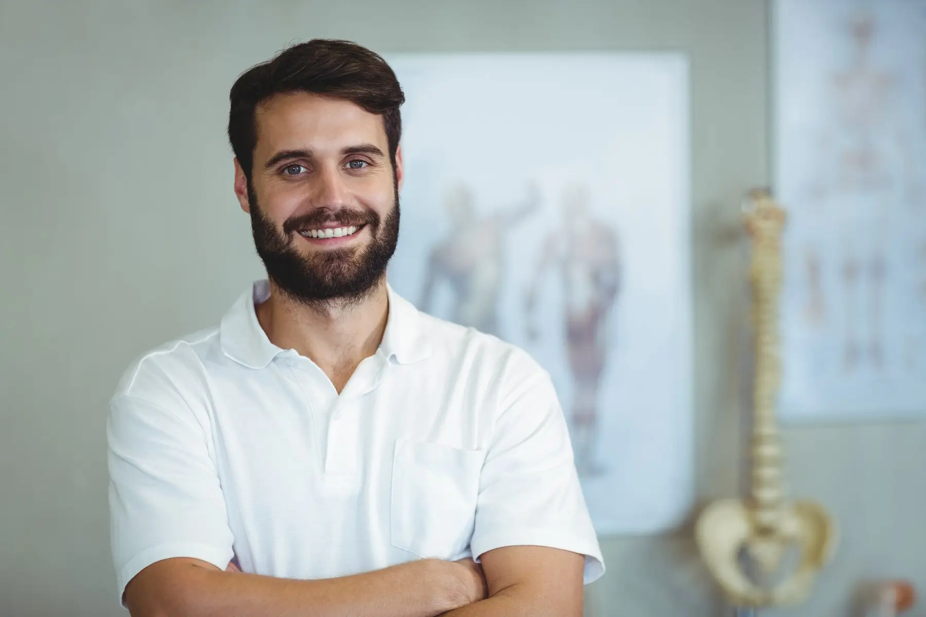 Portrait d’un praticien souriant en polo blanc, bras croisés, devant des planches d’anatomie et un modèle de colonne vertébrale