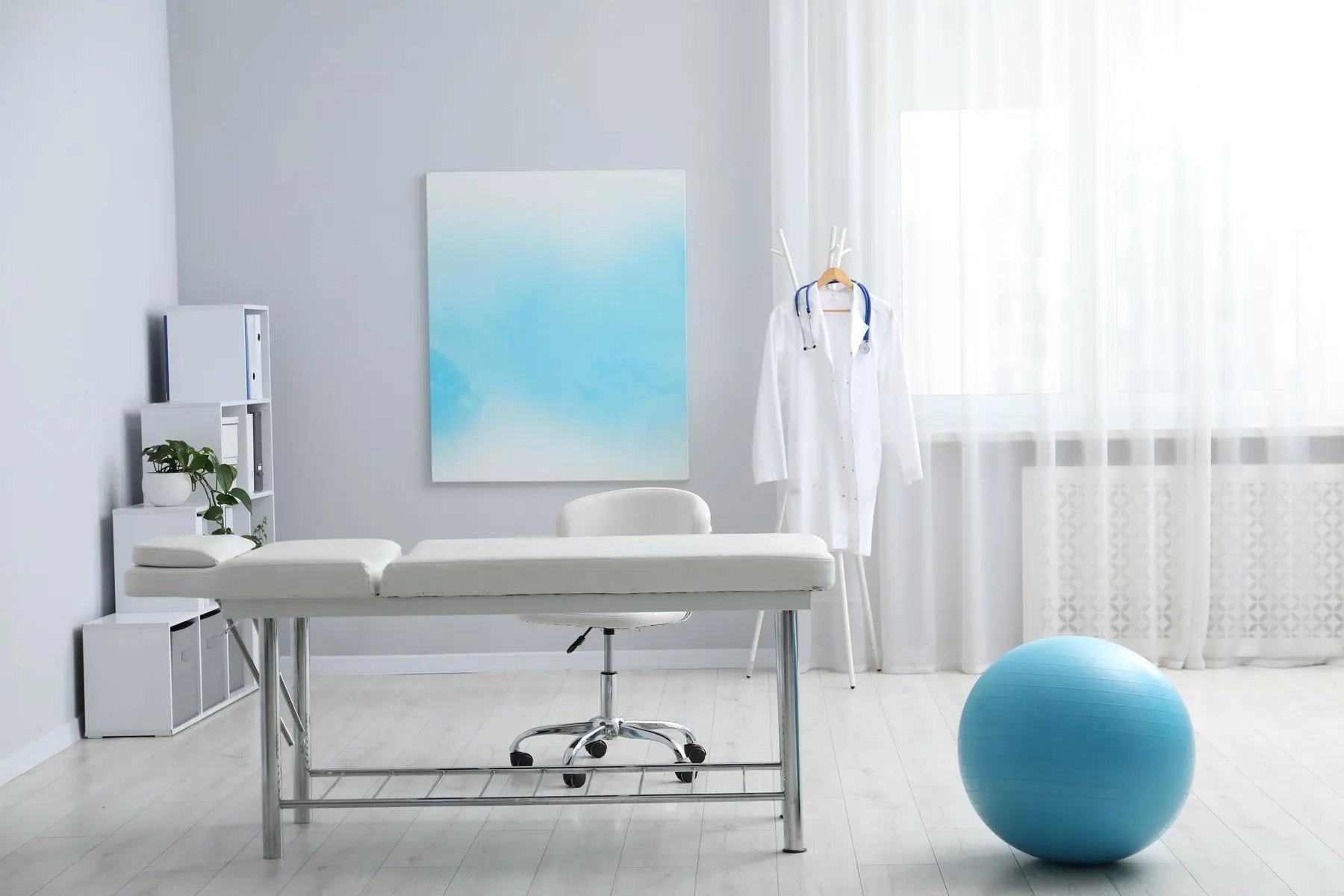 Salle de consultation claire avec une table de soin, un ballon de gymnastique bleu et une blouse blanche sur un portant