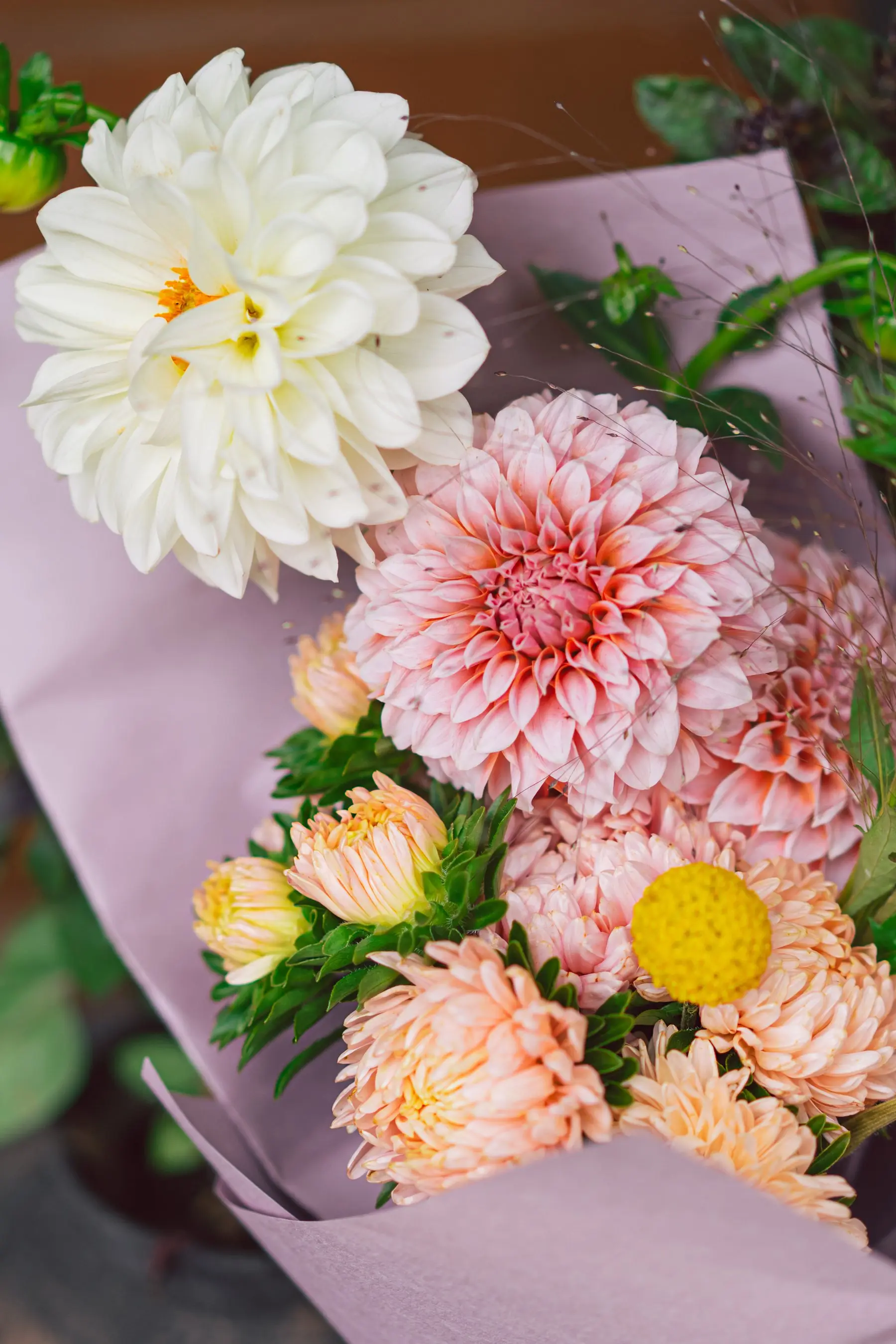 Bouquet de dahlias blancs et rose pêche, d’asters saumon et d’une boule jaune de craspedia, emballé dans un papier mauve