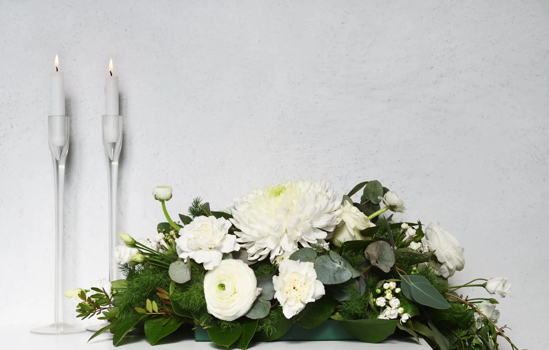 Composition basse de fleurs blanches et de feuillage, deux bougies allumées dans des bougeoirs de verre à côté