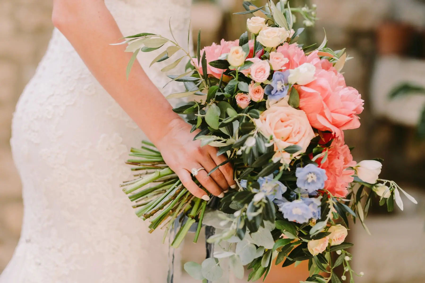 Une mariée en robe de dentelle tient un bouquet de pivoines corail, de roses pêche, de delphiniums bleus et de branches d’olivier
