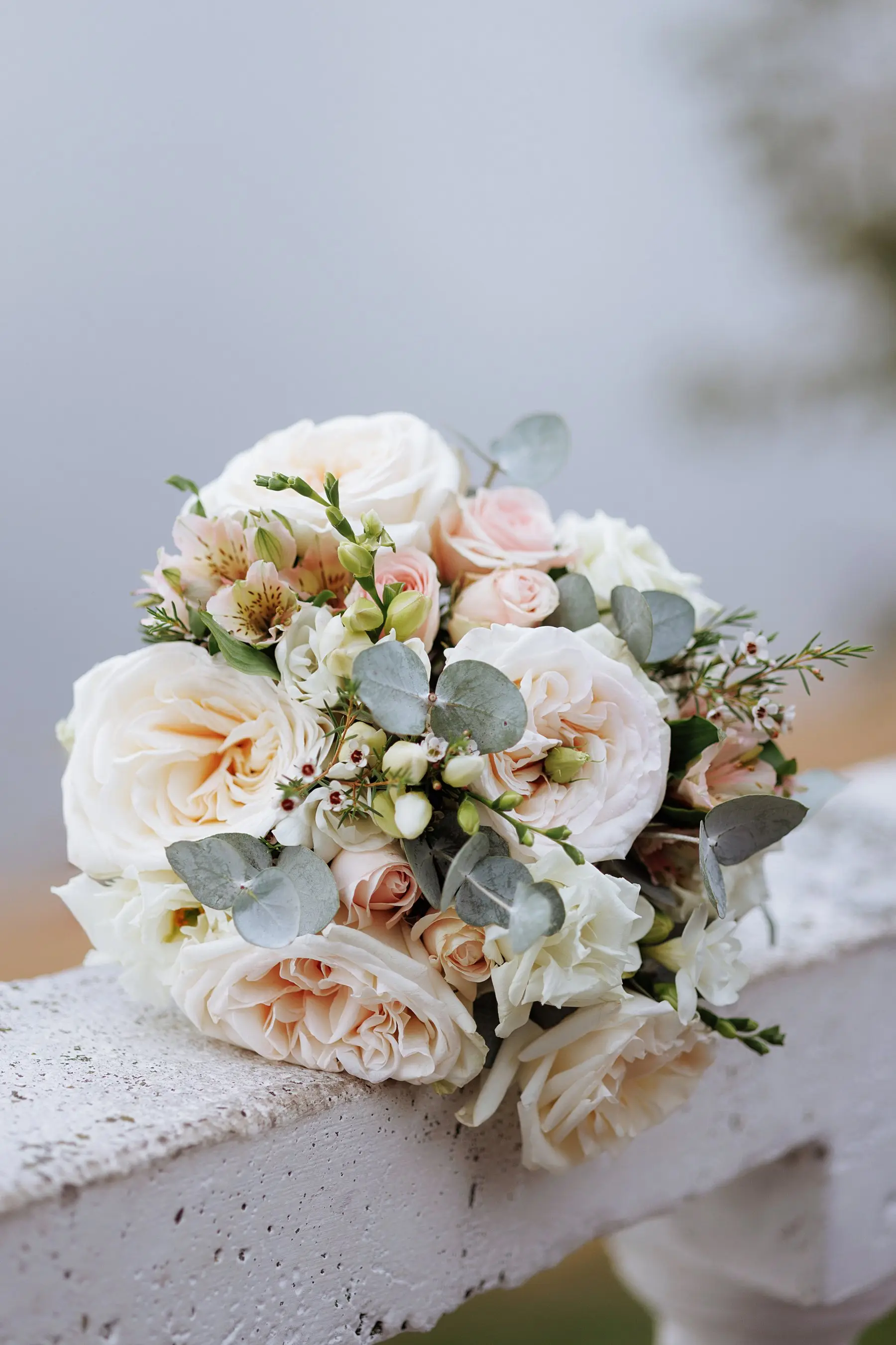 Bouquet rond de roses ivoire et pêche, d’alstroemères et d’eucalyptus posé sur une balustrade de pierre blanche