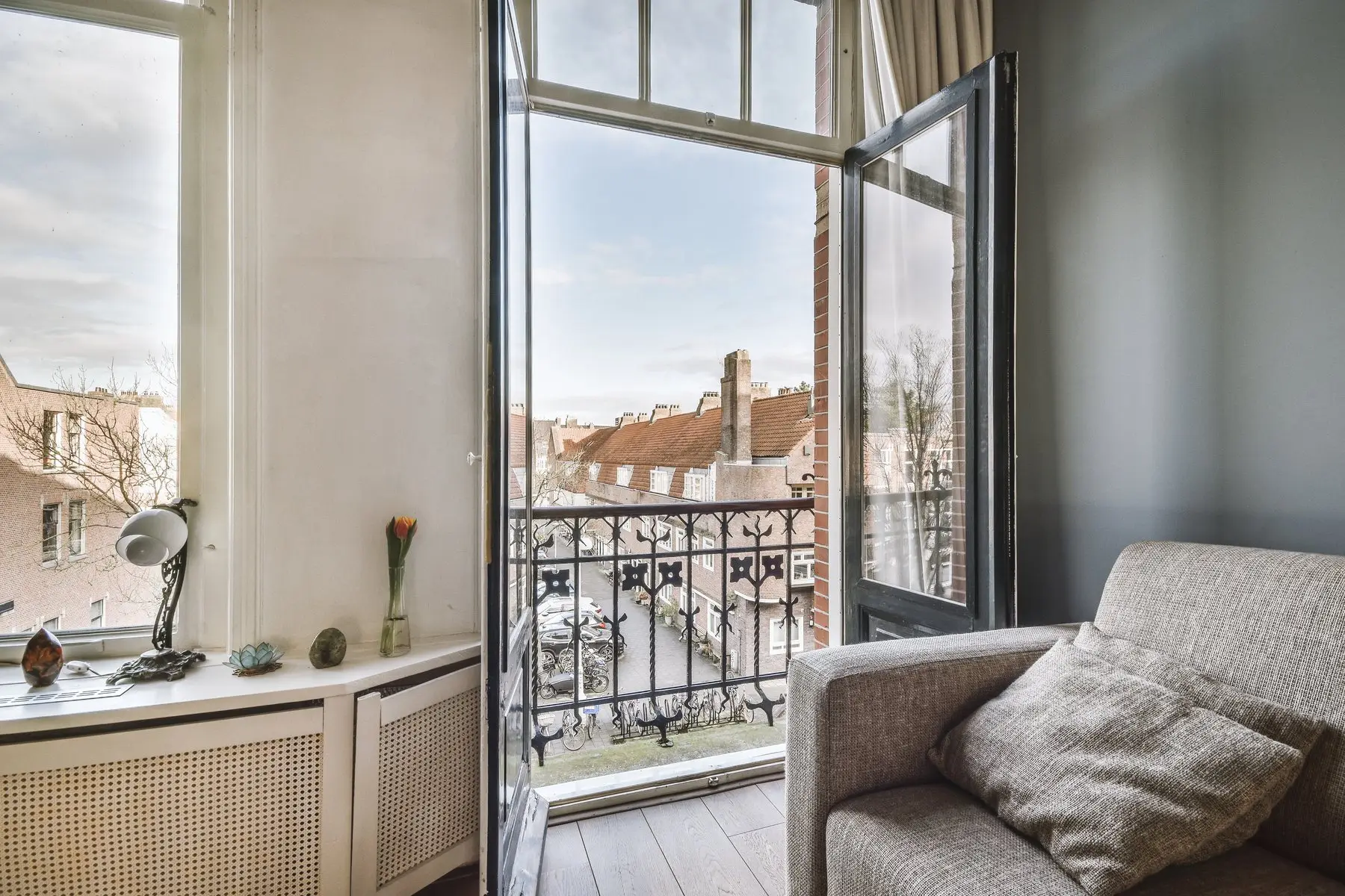 Porte-fenêtre ouverte sur un balcon en fer forgé, vue sur une rue de maisons en brique, fauteuil gris au premier plan